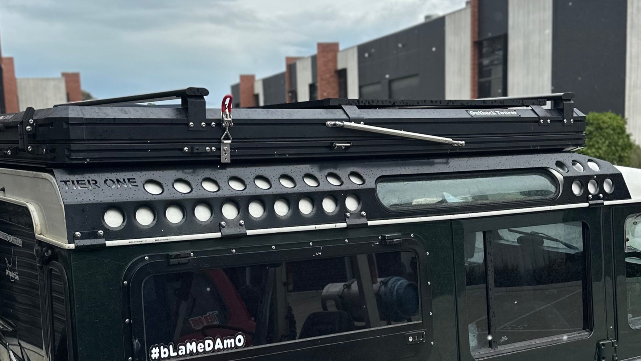 This is an image of a green 4x4 vehicle with a Tier One Metalworks roof rack and an Outback Tent rooftop tent on a rainy day