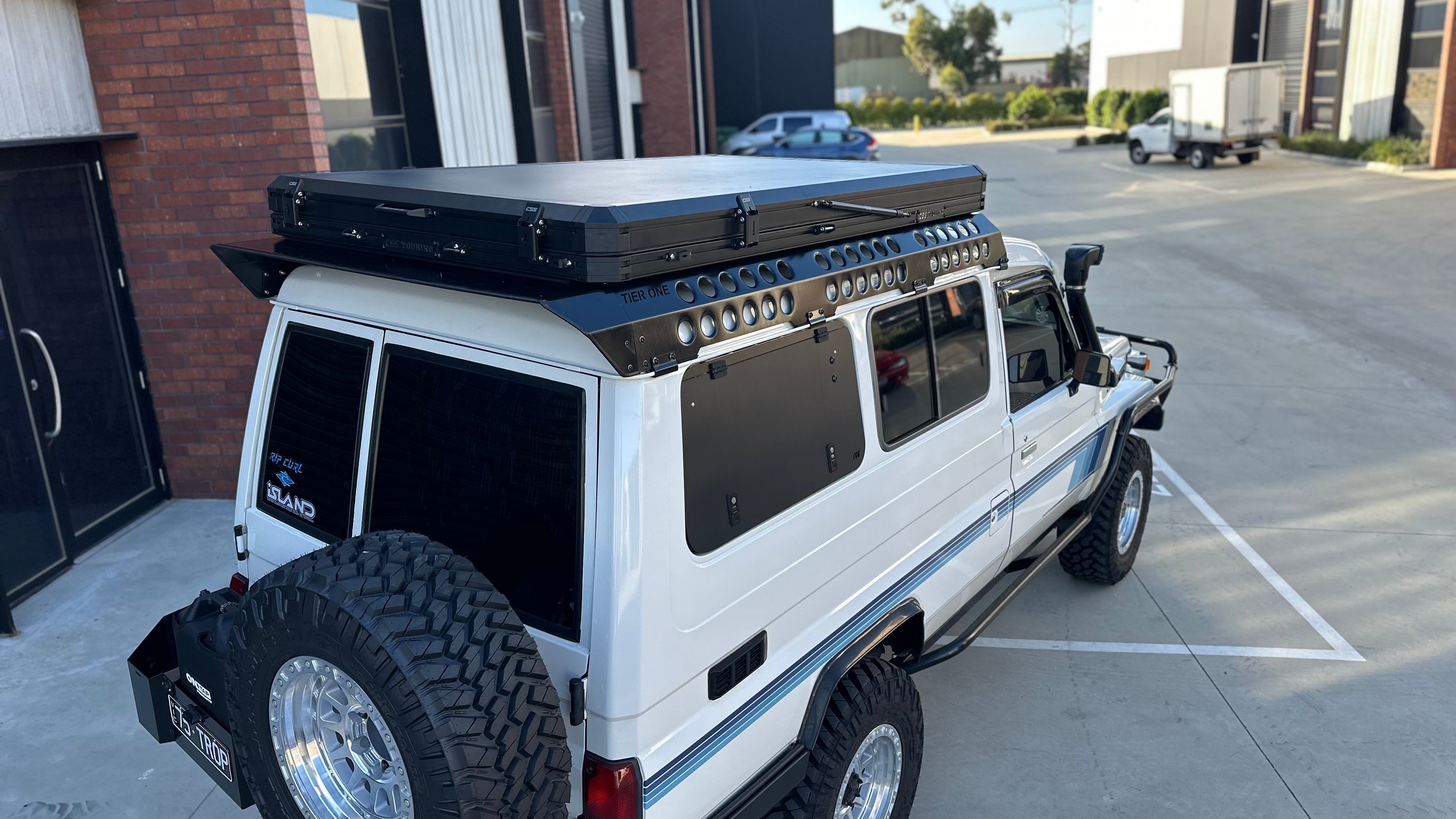 This is an image of a white Toyota Land Cruiser fitted with Tier One Metalworks roof racks and rails, featuring a black rooftop tent in an industrial setting.