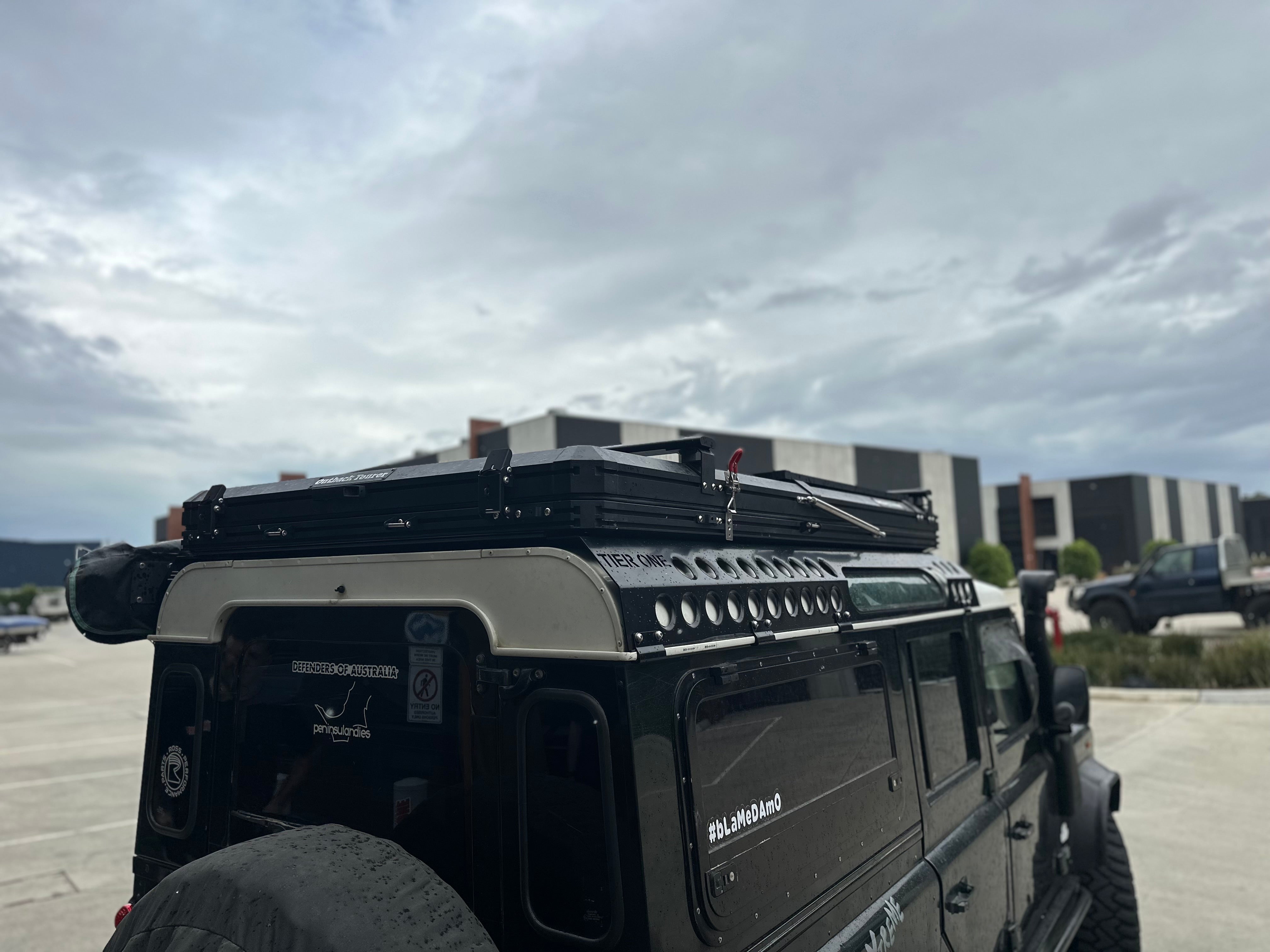 This is an image of a black Land Rover Defender fitted with a Tier One Metalworks roof rack and rails in an outdoor industrial setting