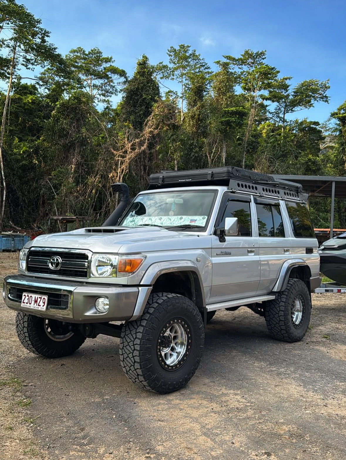 This is an image of a silver Toyota Land Cruiser 4WD fitted with a black Tier One Metalworks roof rack and snorkel, parked on dirt with forest background
