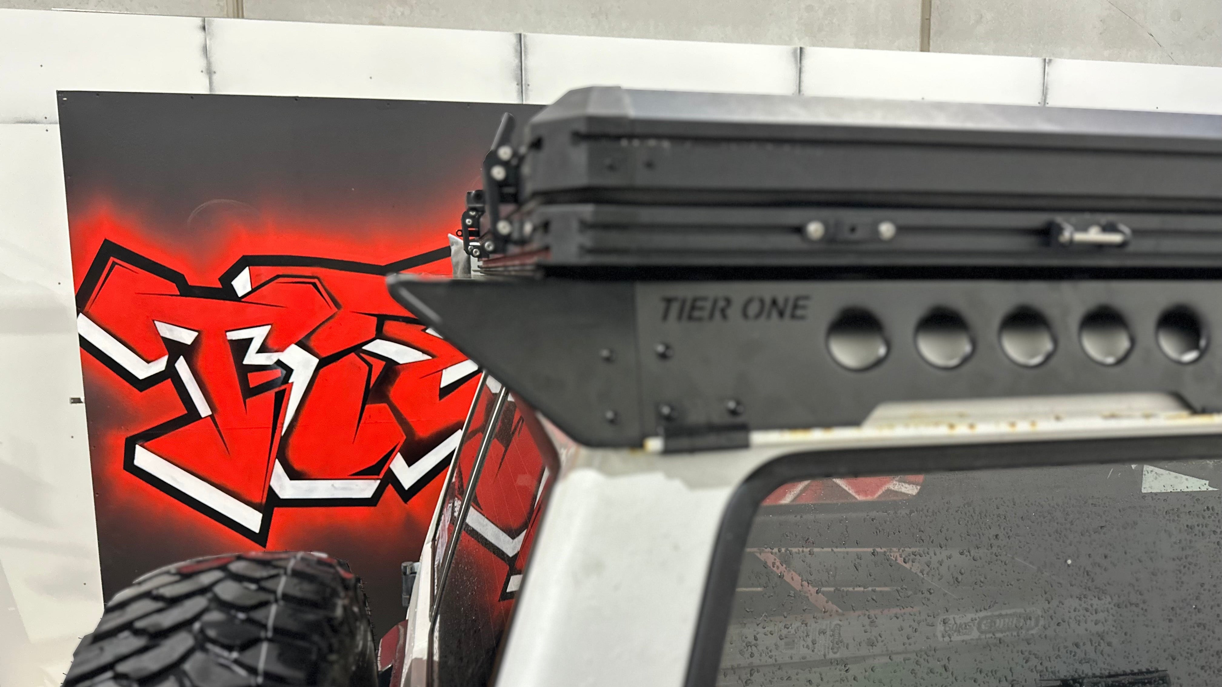 This is an image of a white 4x4 with a black Tier One Metalworks roof rack mounted, photographed inside a workshop with graffiti art in the background.