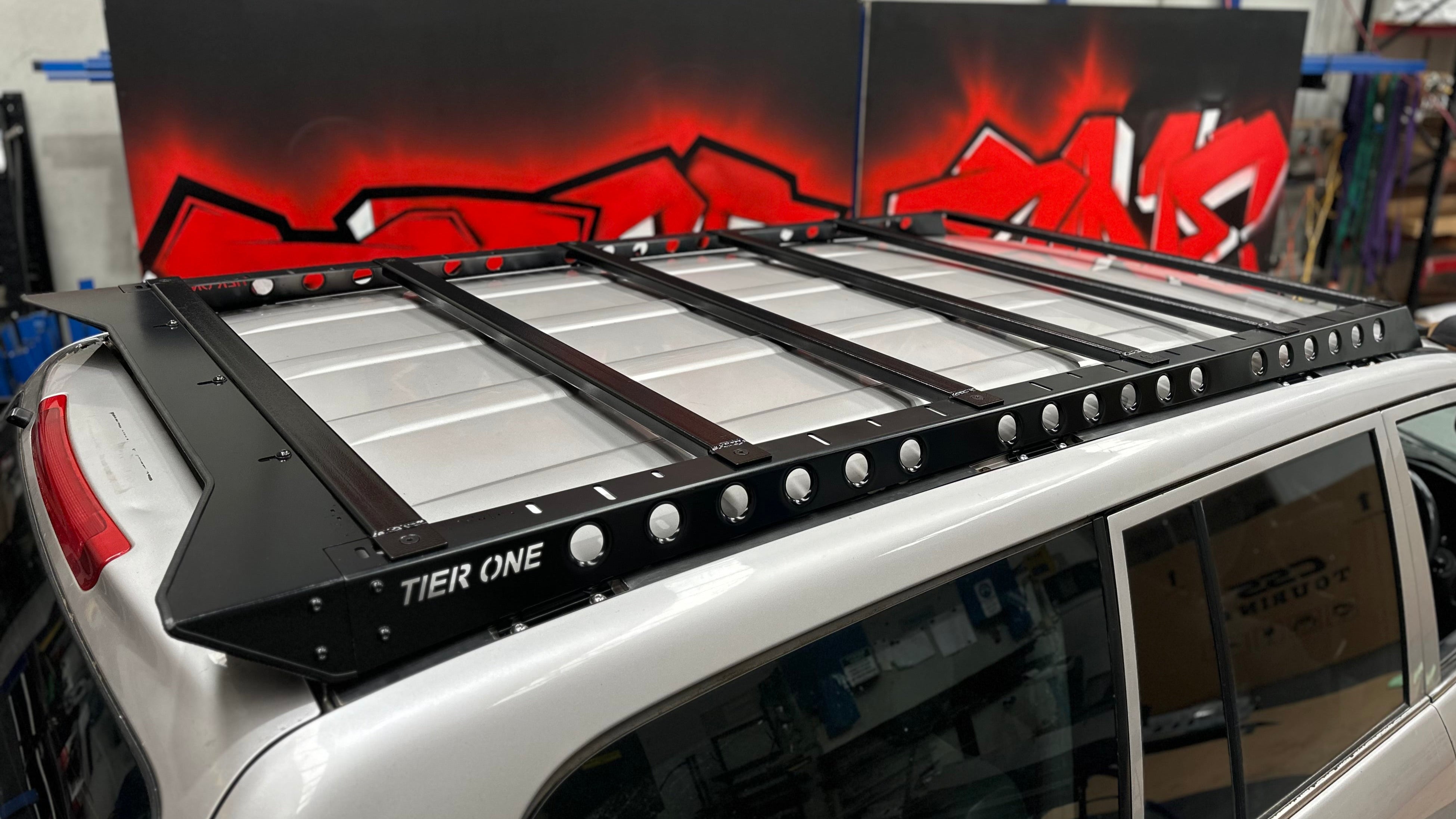 This is an image of a silver 4WD vehicle fitted with a black Tier One Metalworks roof rack inside a workshop