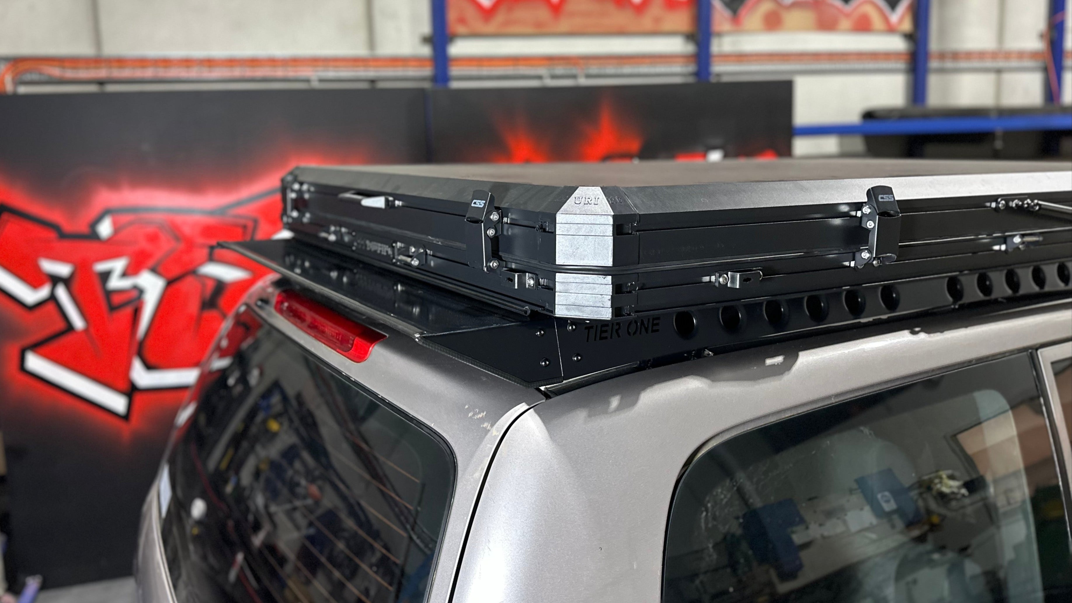 This is an image of a silver 4x4 with a Tier One Metalworks black roof rack and a closed CSS Touring rooftop tent on top