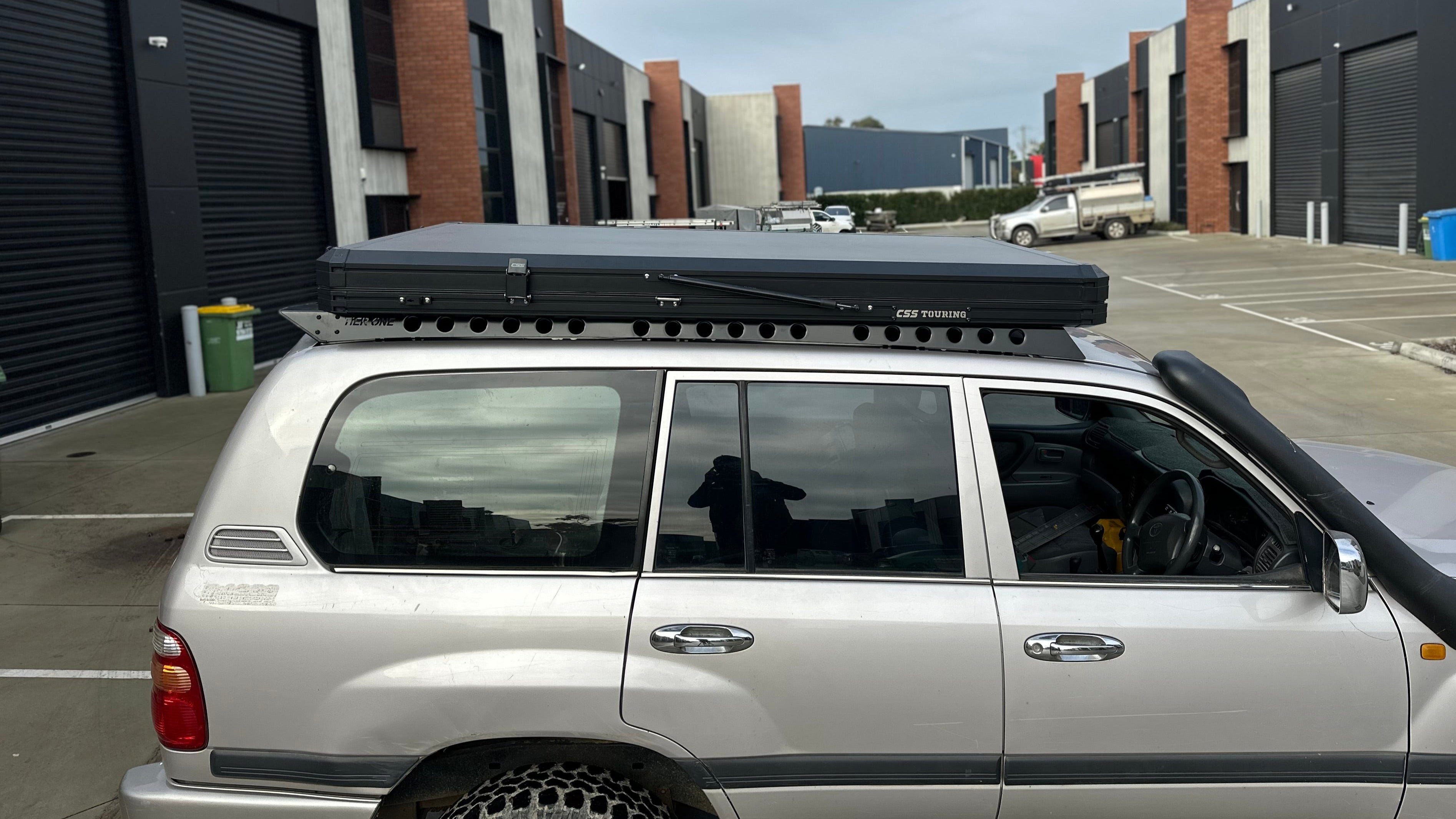 This is an image of a silver Toyota Land Cruiser with a Tier One Metalworks roof rack and CSS Touring rooftop tent in an industrial parking area