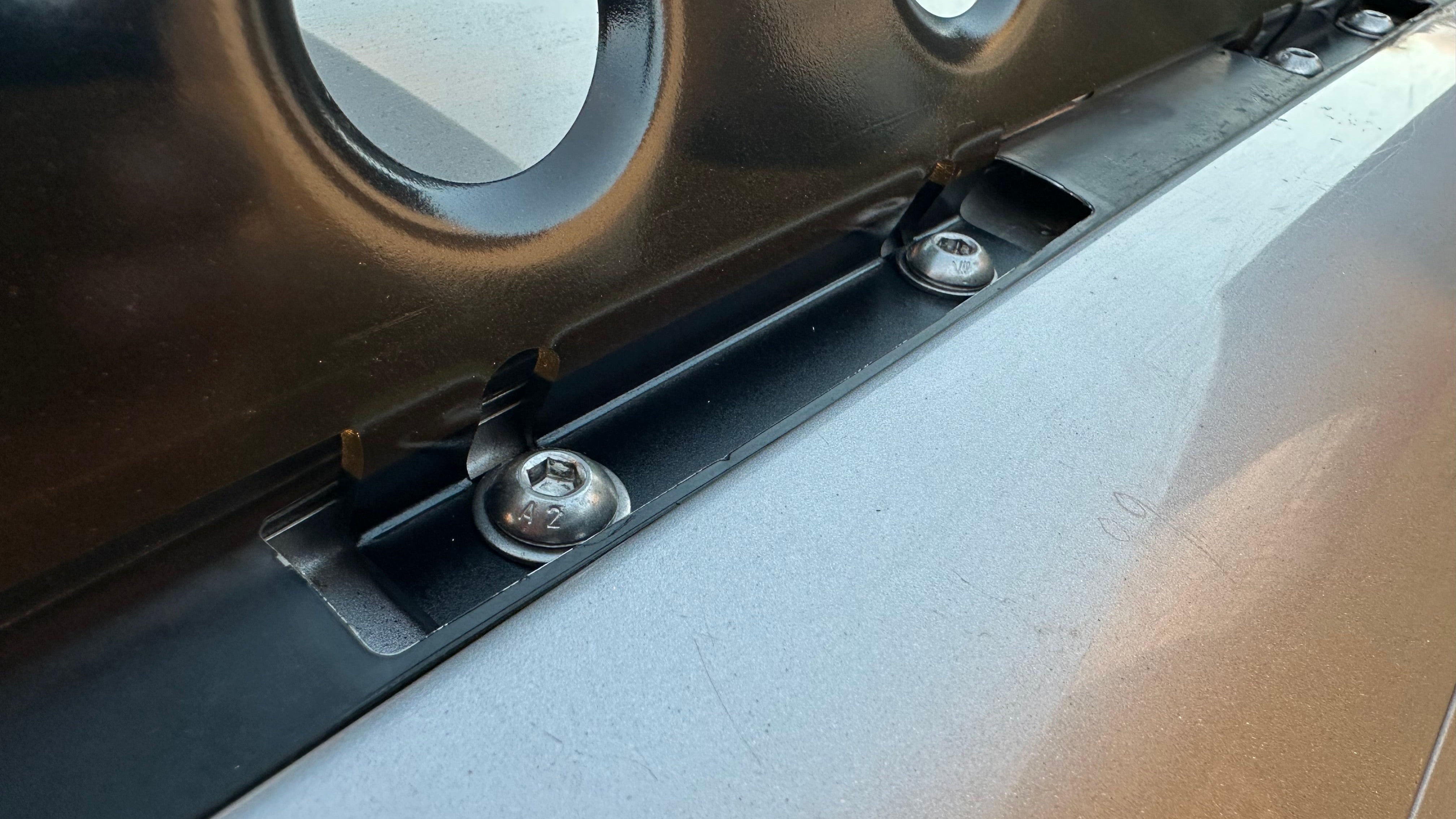 This is an image of a close-up view of bolts fastening a black metal component to a silver surface, likely part of Tier One Metalworks roof rack assembly.