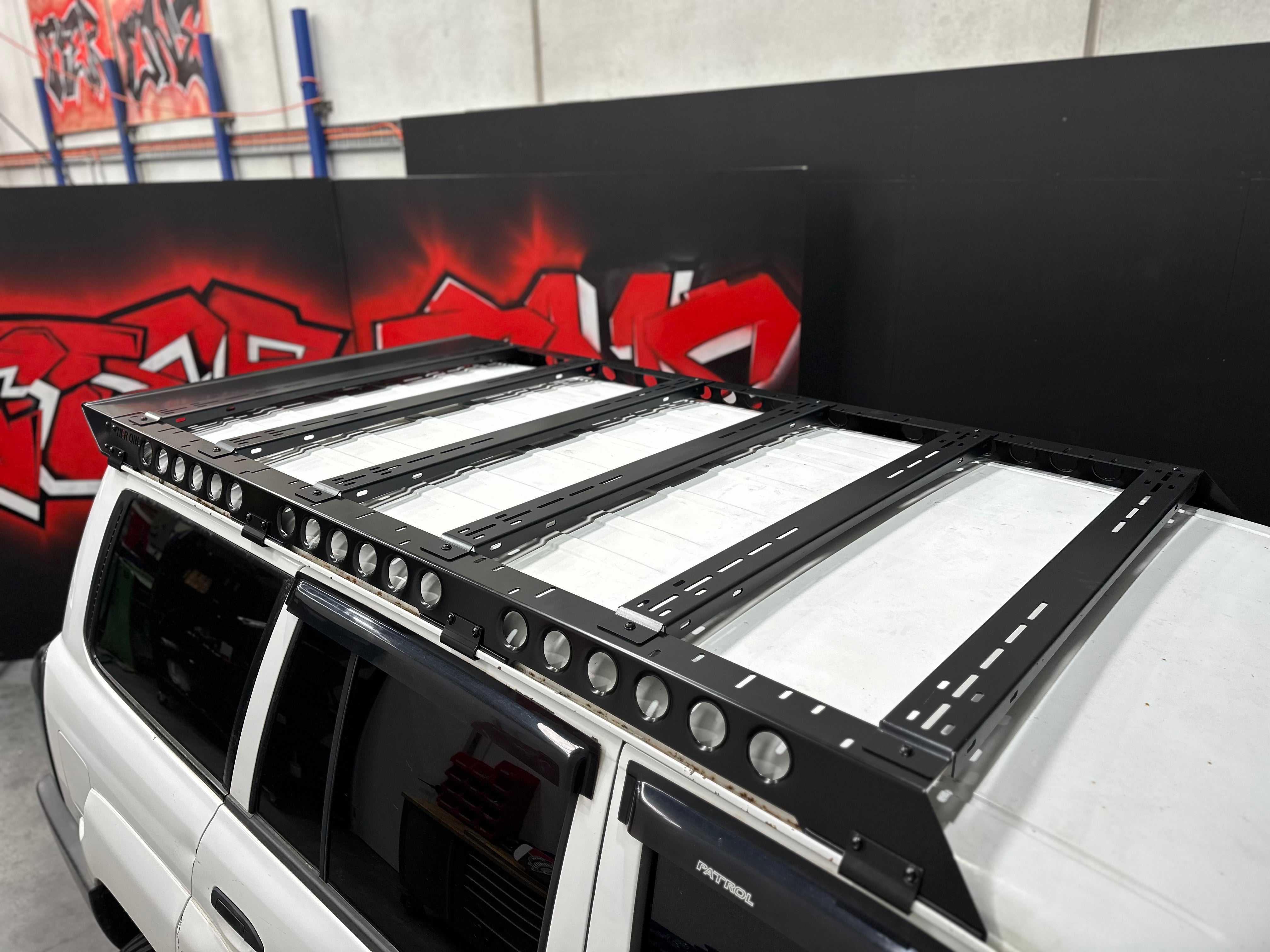 This is an image of a white Nissan Patrol fitted with a black Tier One Metalworks Roof Rack and Rails inside a workshop with graffiti art