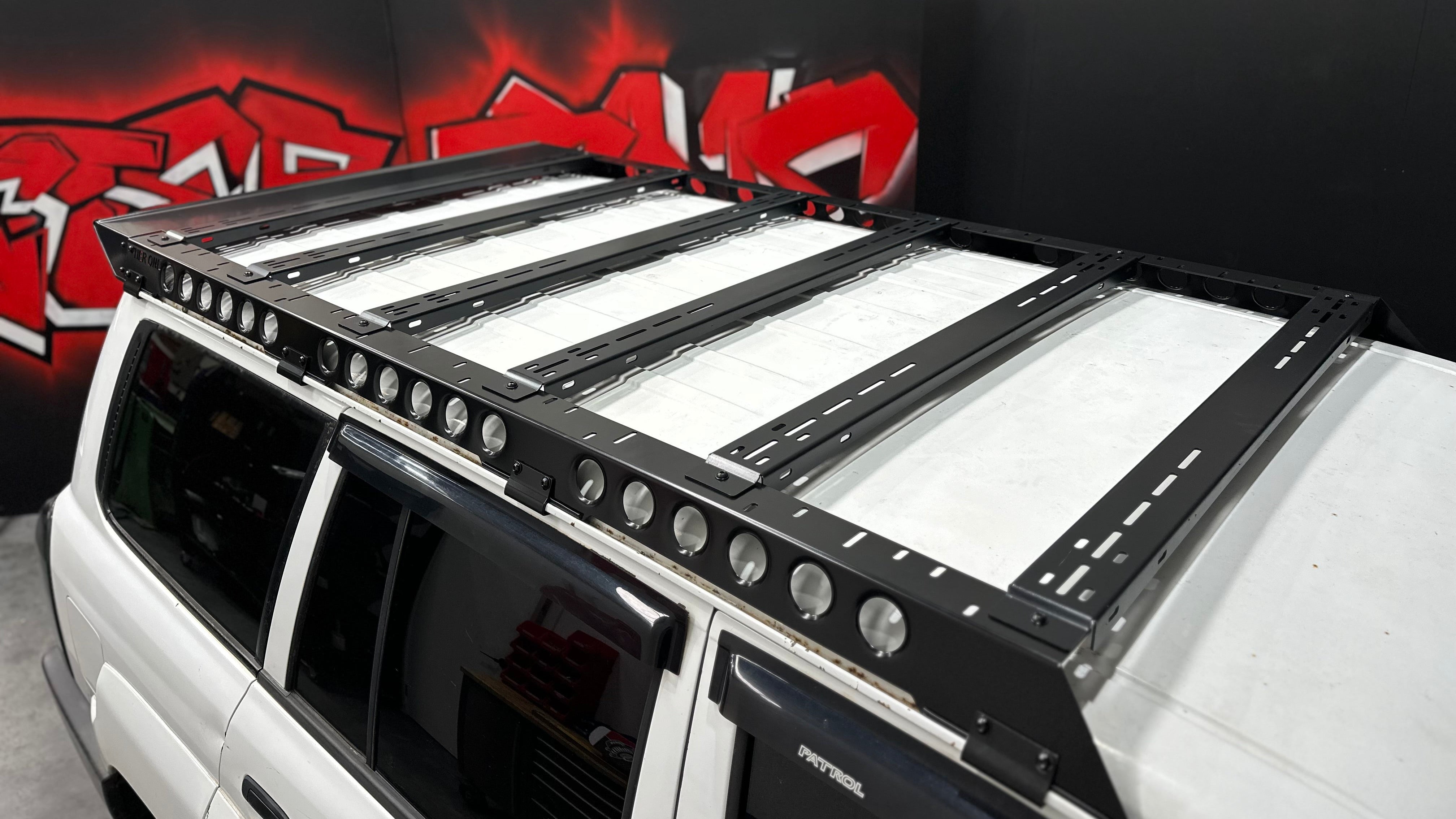 This is an image of a white Nissan Patrol fitted with a black Tier One Metalworks Roof Rack and Rails inside a workshop with graffiti art