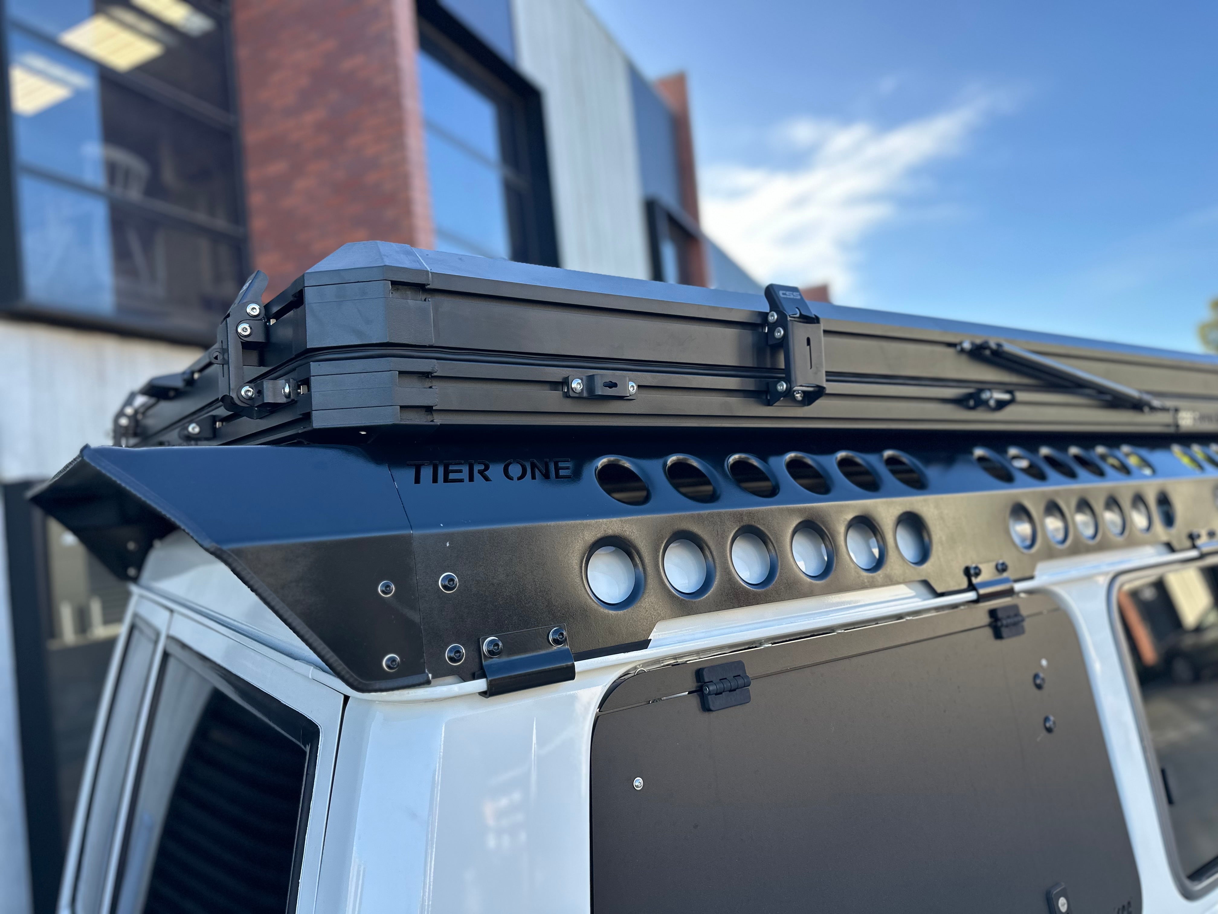 This is an image of a white 4x4 vehicle fitted with black Tier One Metalworks Roof Racks and Rails, parked outside a building.