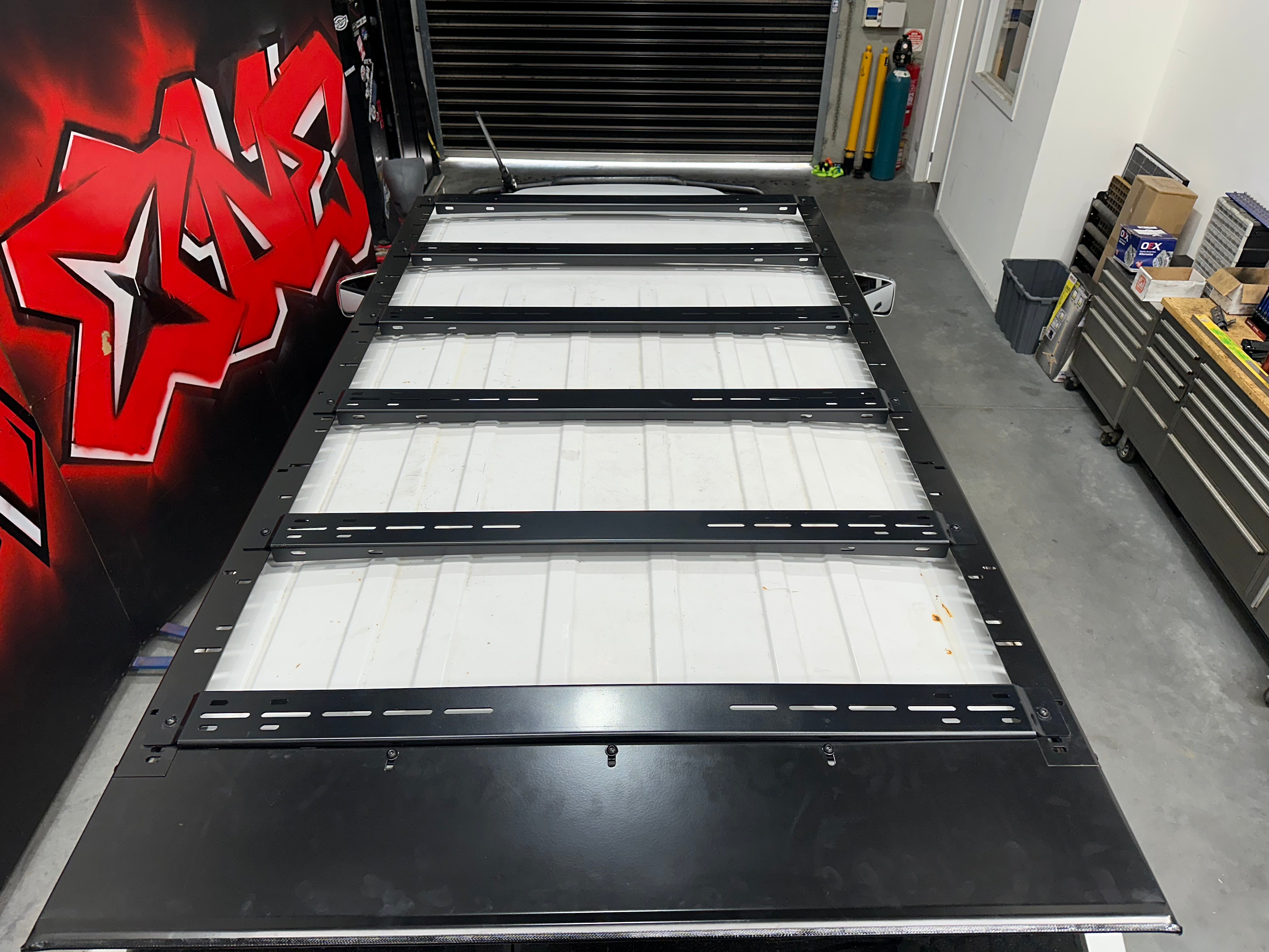 This is an image of a black Tier One Metalworks steel roof rack mounted on a white vehicle tray inside a workshop
