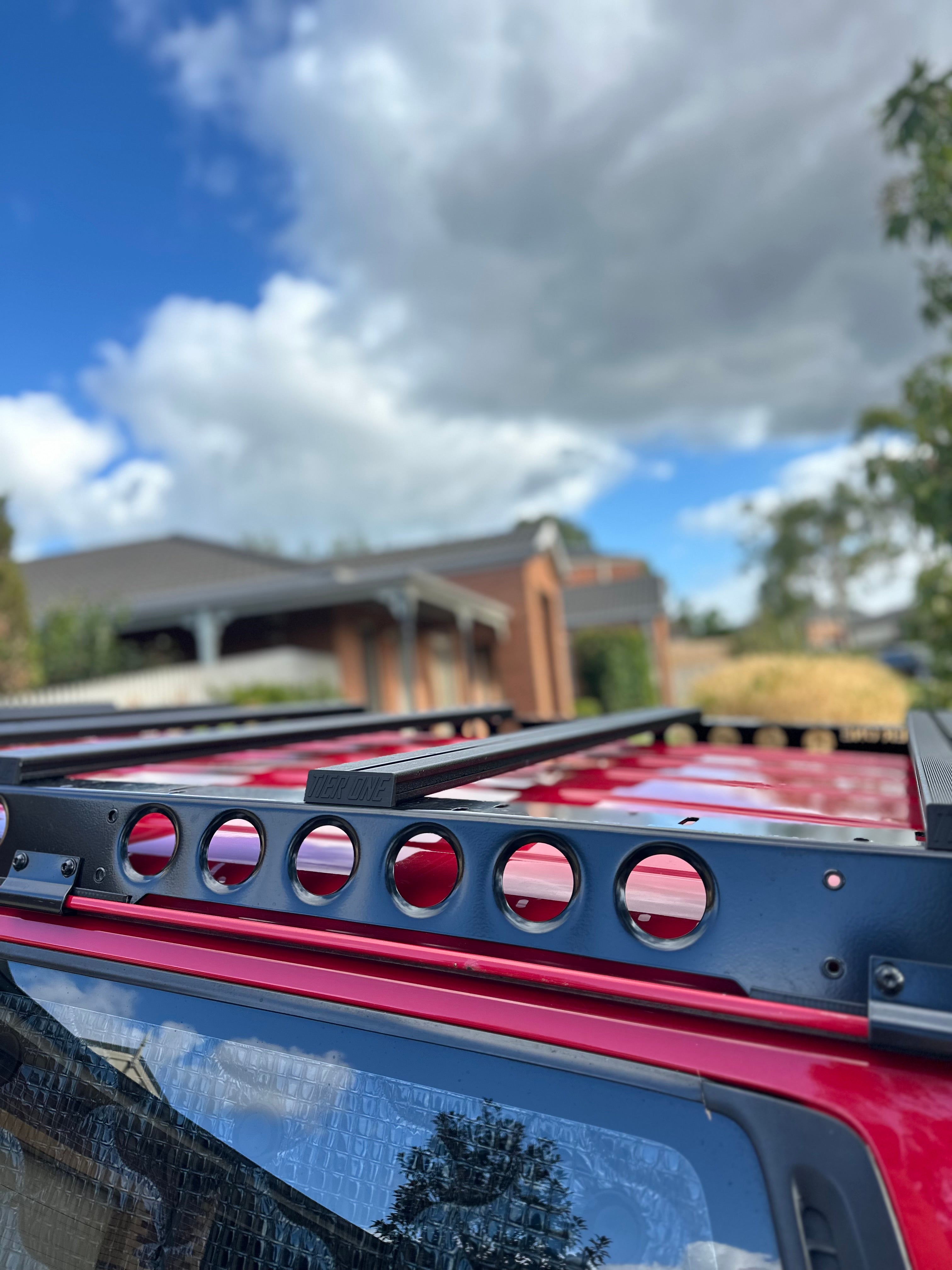This is an image of a black Tier One Metalworks Roof Rack mounted on a red 4x4 vehicle roof under a partly cloudy sky