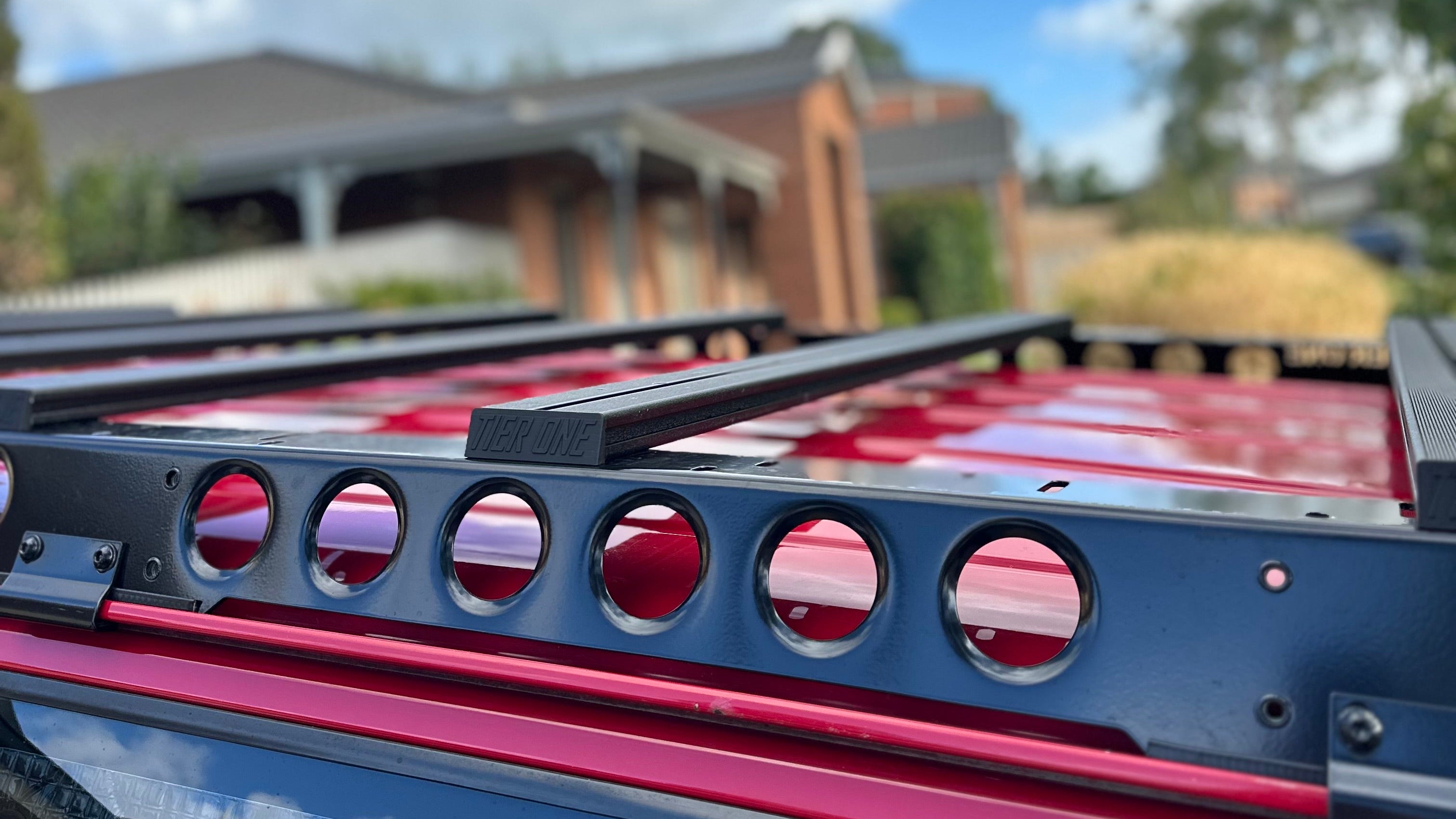 This is an image of a black Tier One Metalworks Roof Rack mounted on a red 4x4 vehicle roof under a partly cloudy sky