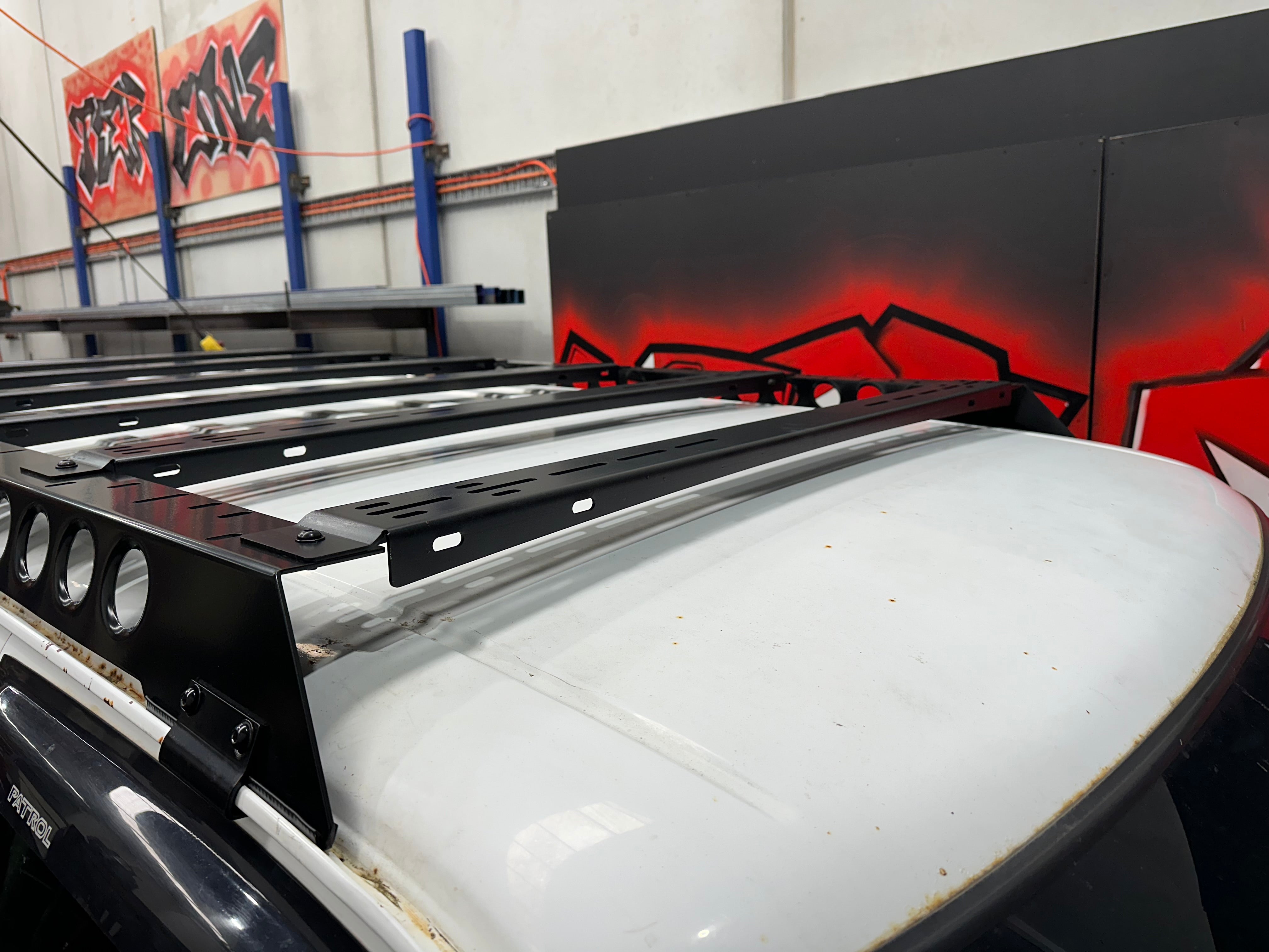 This is an image of a Tier One Metalworks black steel roof rack mounted on a white Nissan Patrol in a workshop