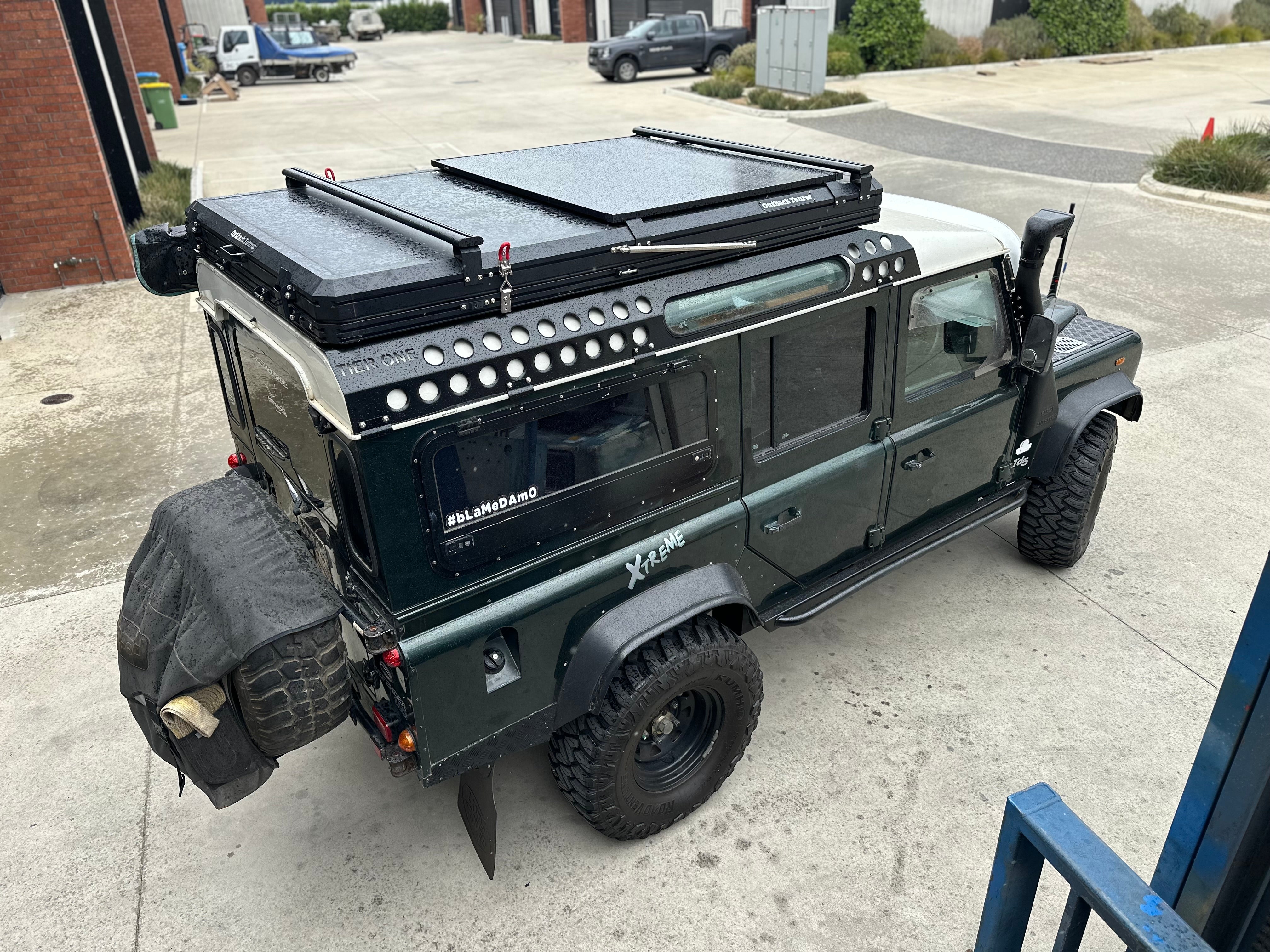 This is an image of a green Land Rover Defender fitted with Tier One Metalworks Roof Racks & Rails and an Outback Terrain roof system in an industrial driveway.