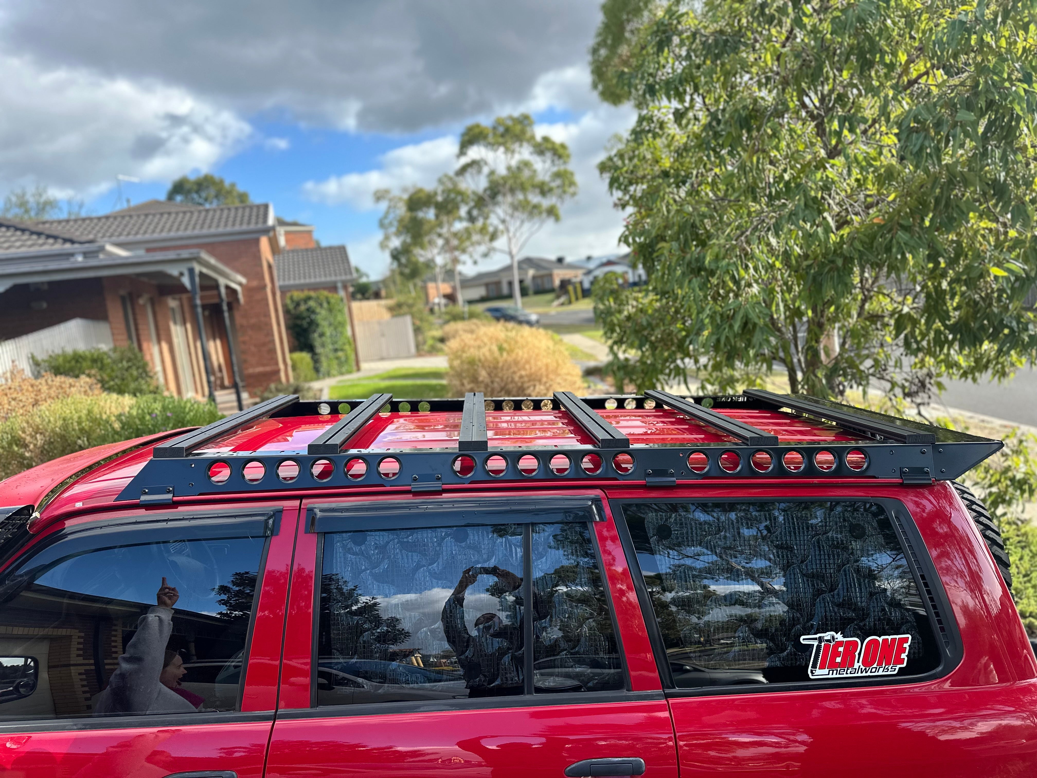 This is an image of a red SUV with a black Tier One Metalworks roof rack installed in a suburban street setting.