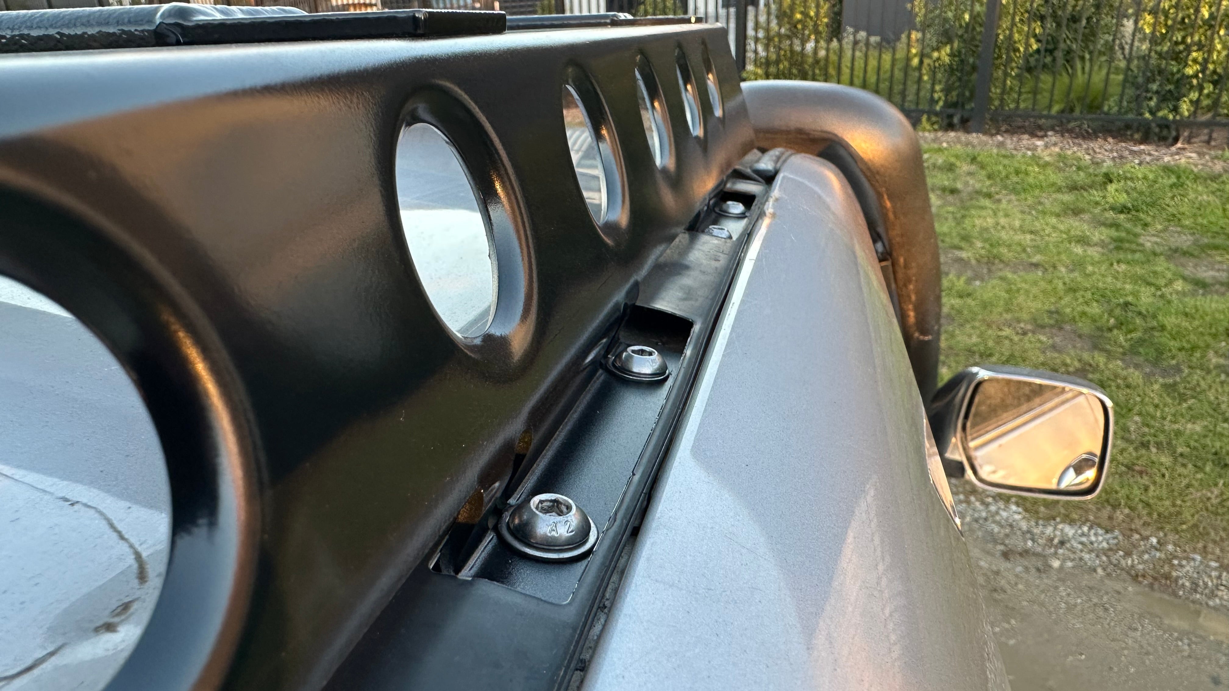 This is an image of a close-up detail of black Tier One Metalworks Roof Rack bolts on a silver 4x4 vehicle with a side mirror and grassy background