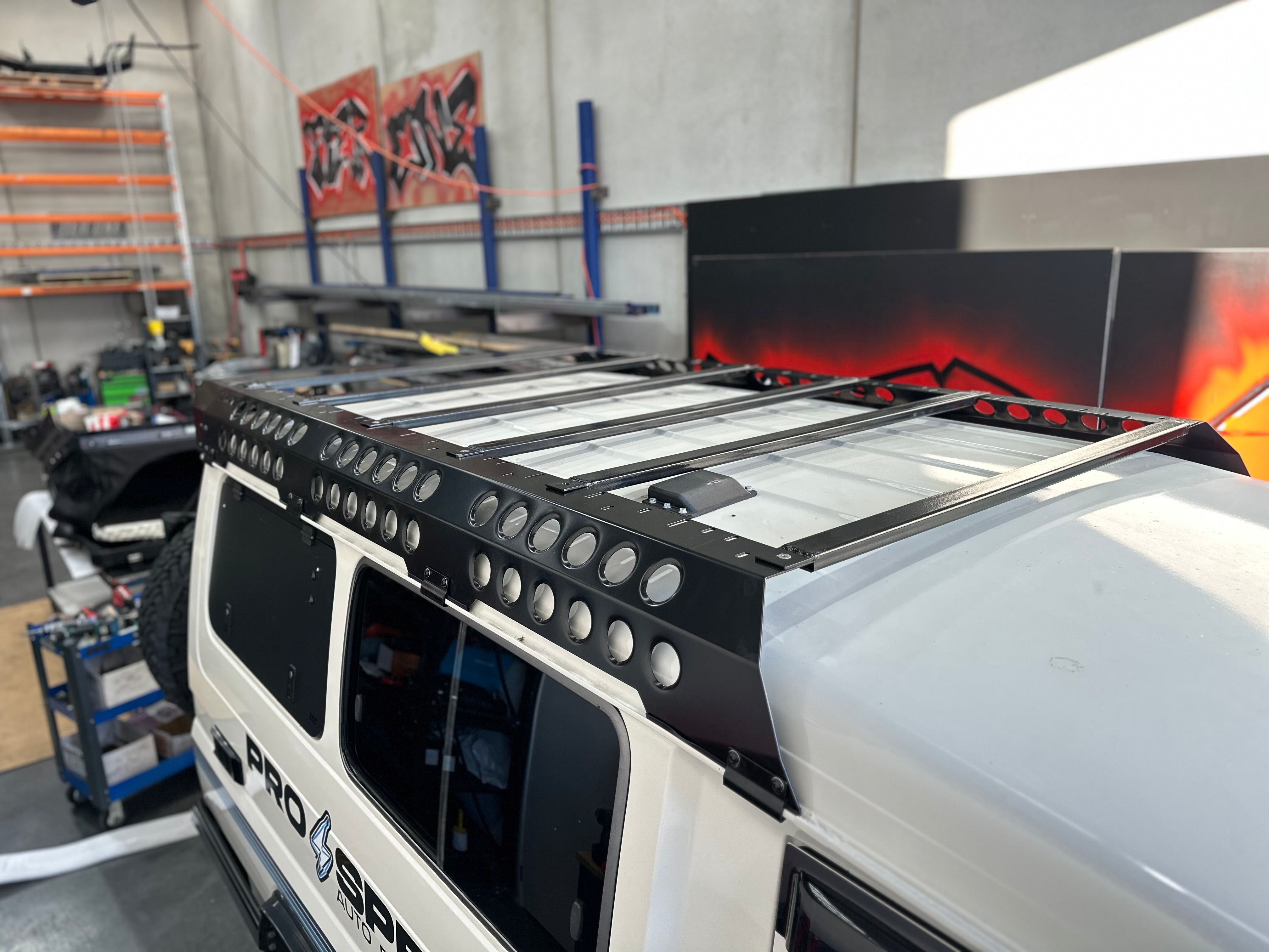 This is an image of a white 4x4 with a black Tier One Metalworks Roof Rack & Rails installed inside a workshop