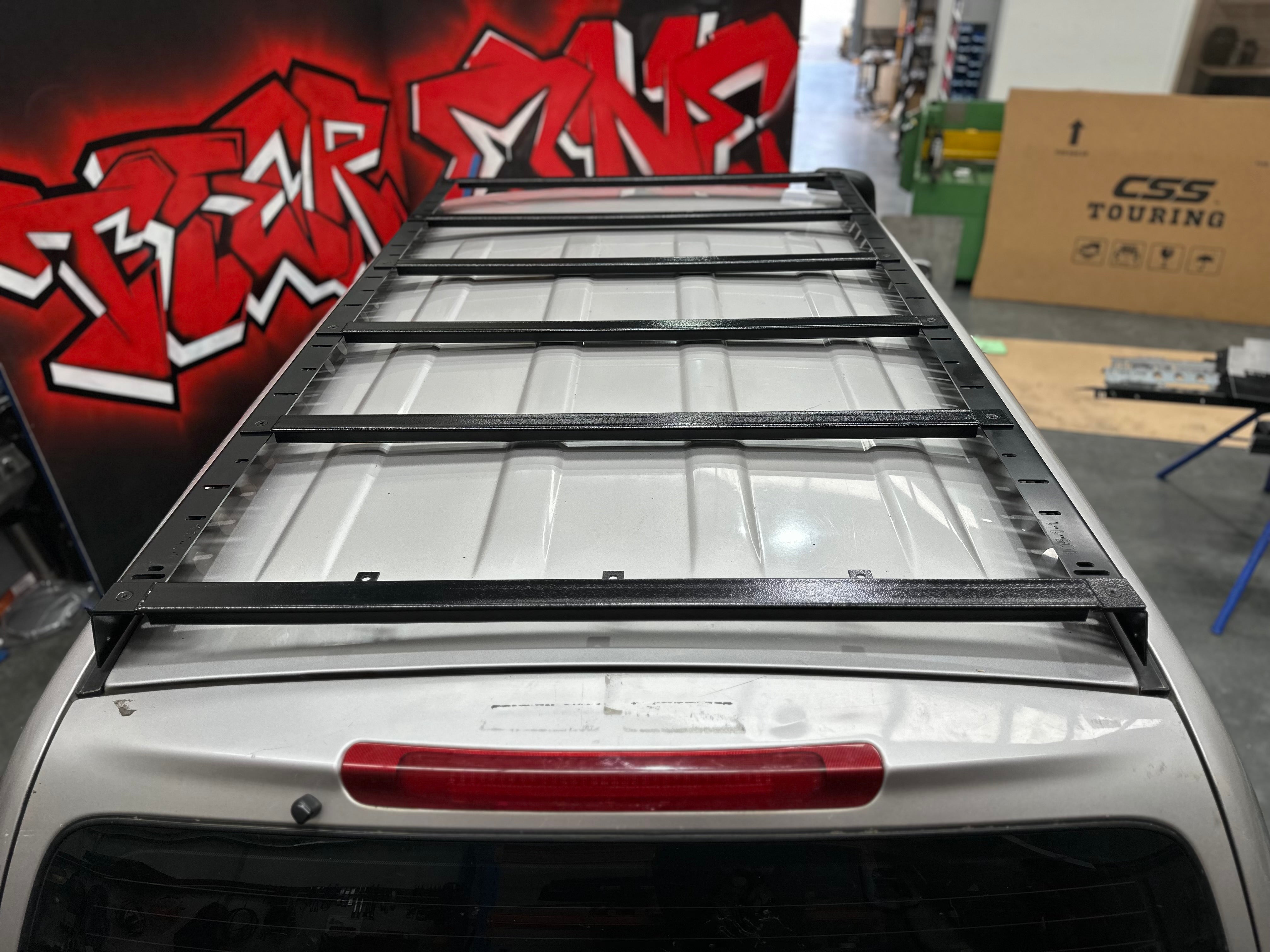 This is an image of a black Tier One Metalworks Roof Rack mounted on a silver 4x4 vehicle roof in a workshop with red graffiti wall.