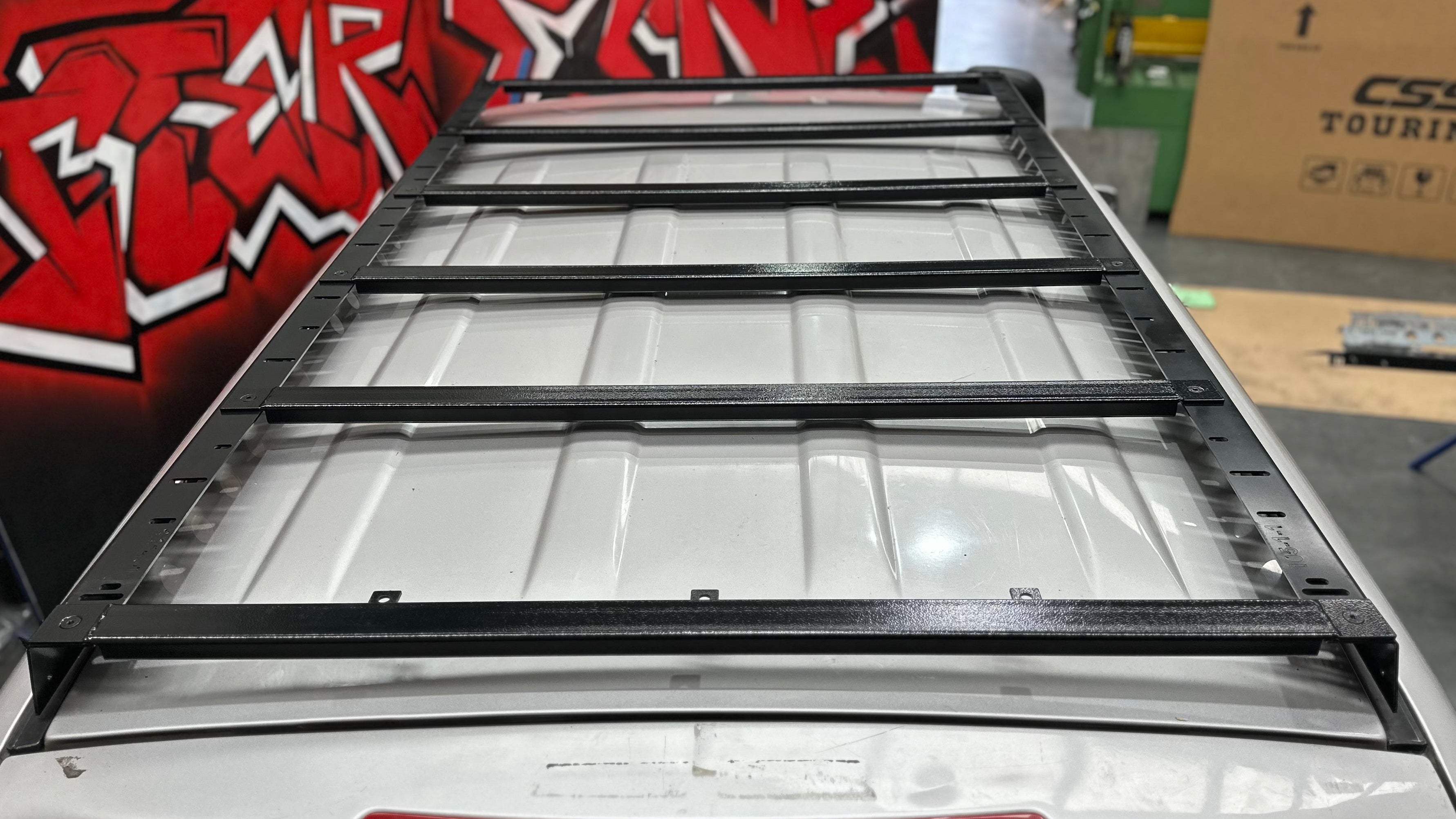 This is an image of a black Tier One Metalworks Roof Rack mounted on a silver 4x4 vehicle roof in a workshop with red graffiti wall.