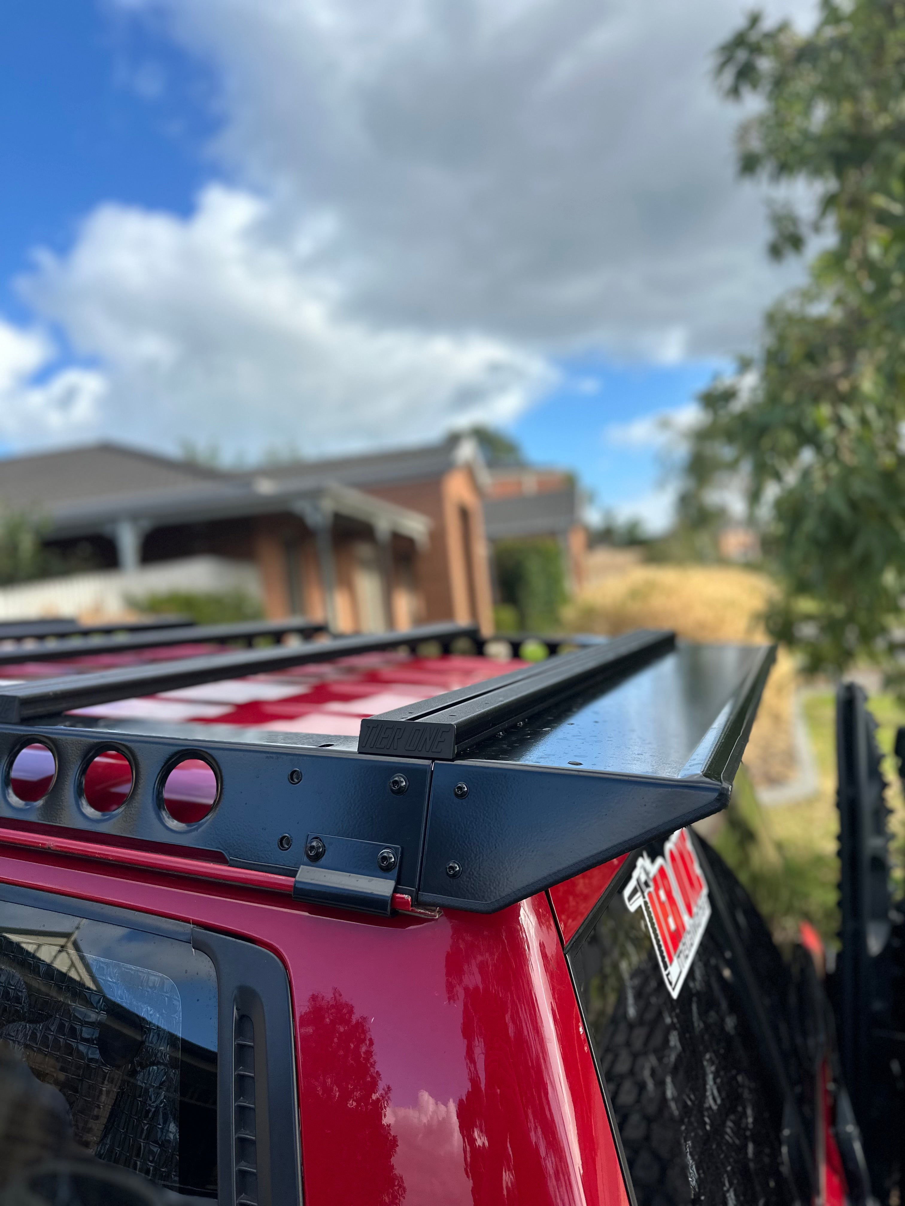 This is an image of a black Tier One Metalworks roof rack mounted on a red 4x4 vehicle roof in a suburban setting