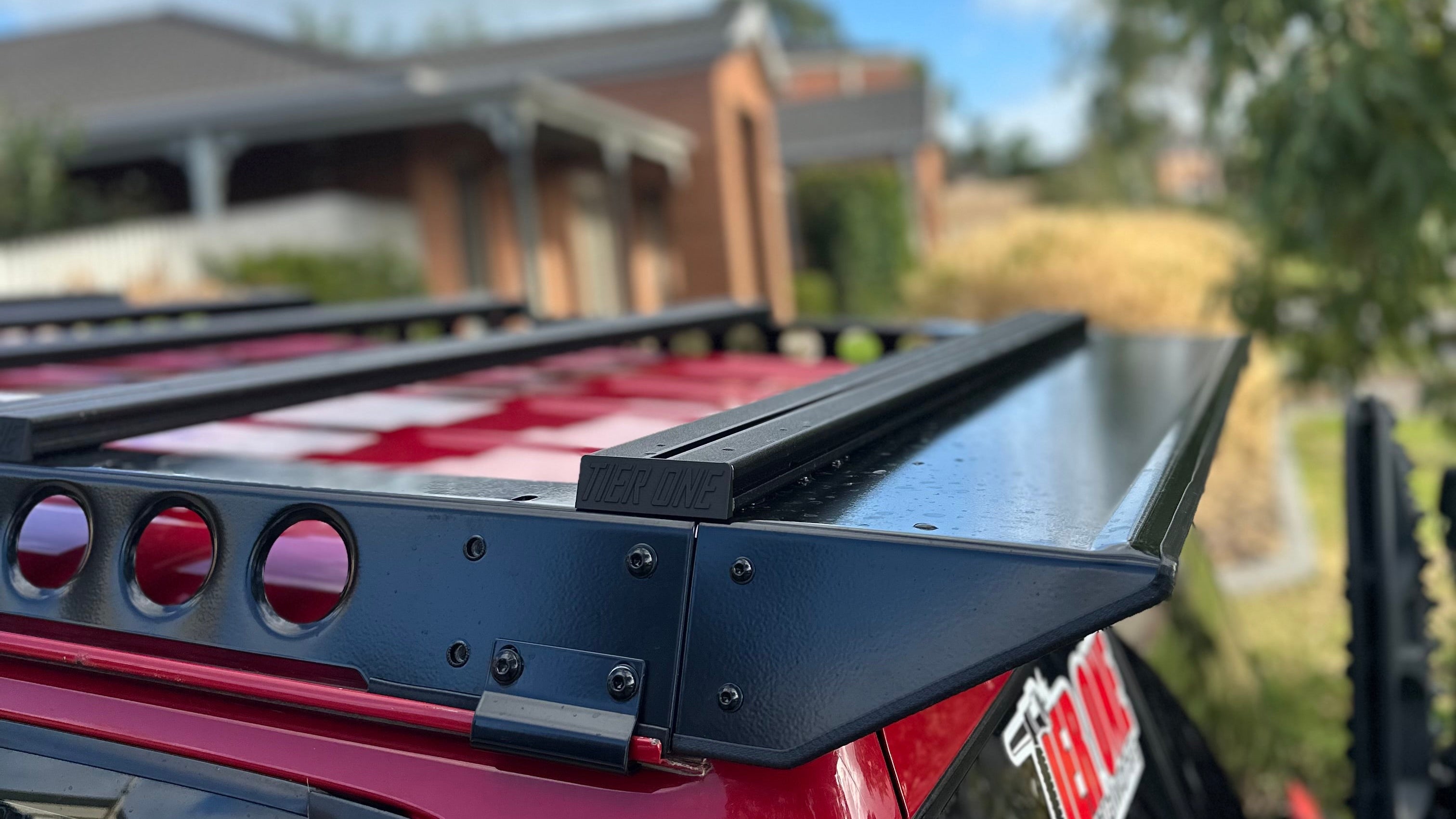 This is an image of a black Tier One Metalworks roof rack mounted on a red 4x4 vehicle roof in a suburban setting