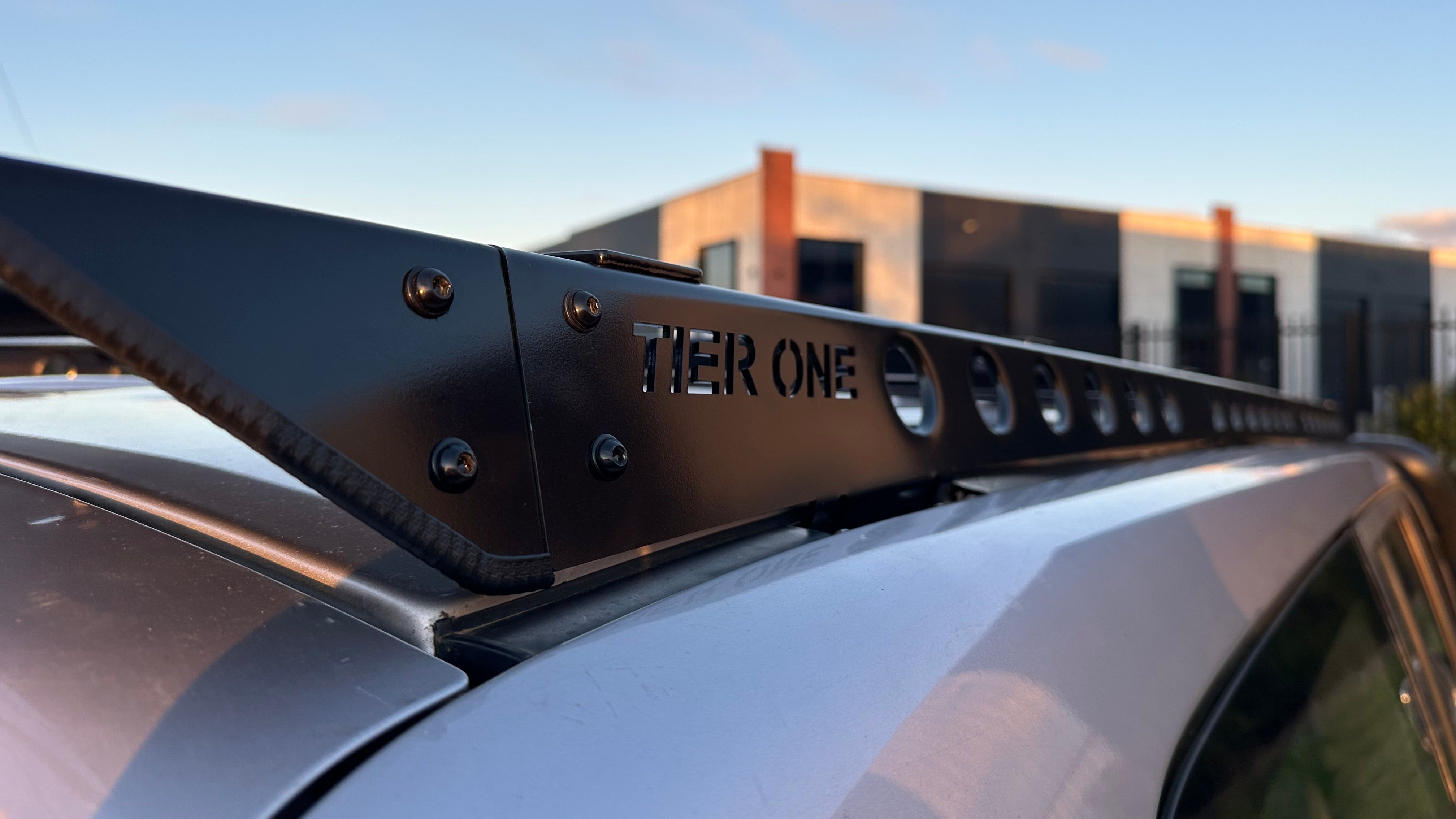 This is an image of a black Tier One Metalworks roof rack mounted on a silver vehicle with a blurred industrial building in the background