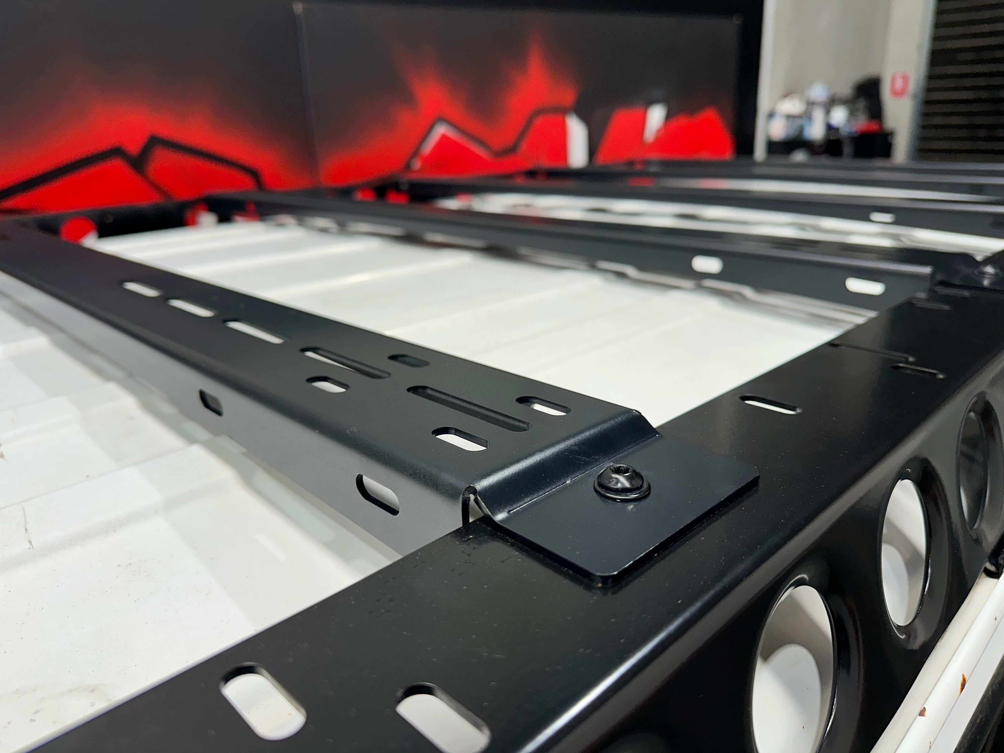 This is an image of a black Tier One Metalworks roof rack with multiple mounting slots on a white vehicle roof inside a workshop
