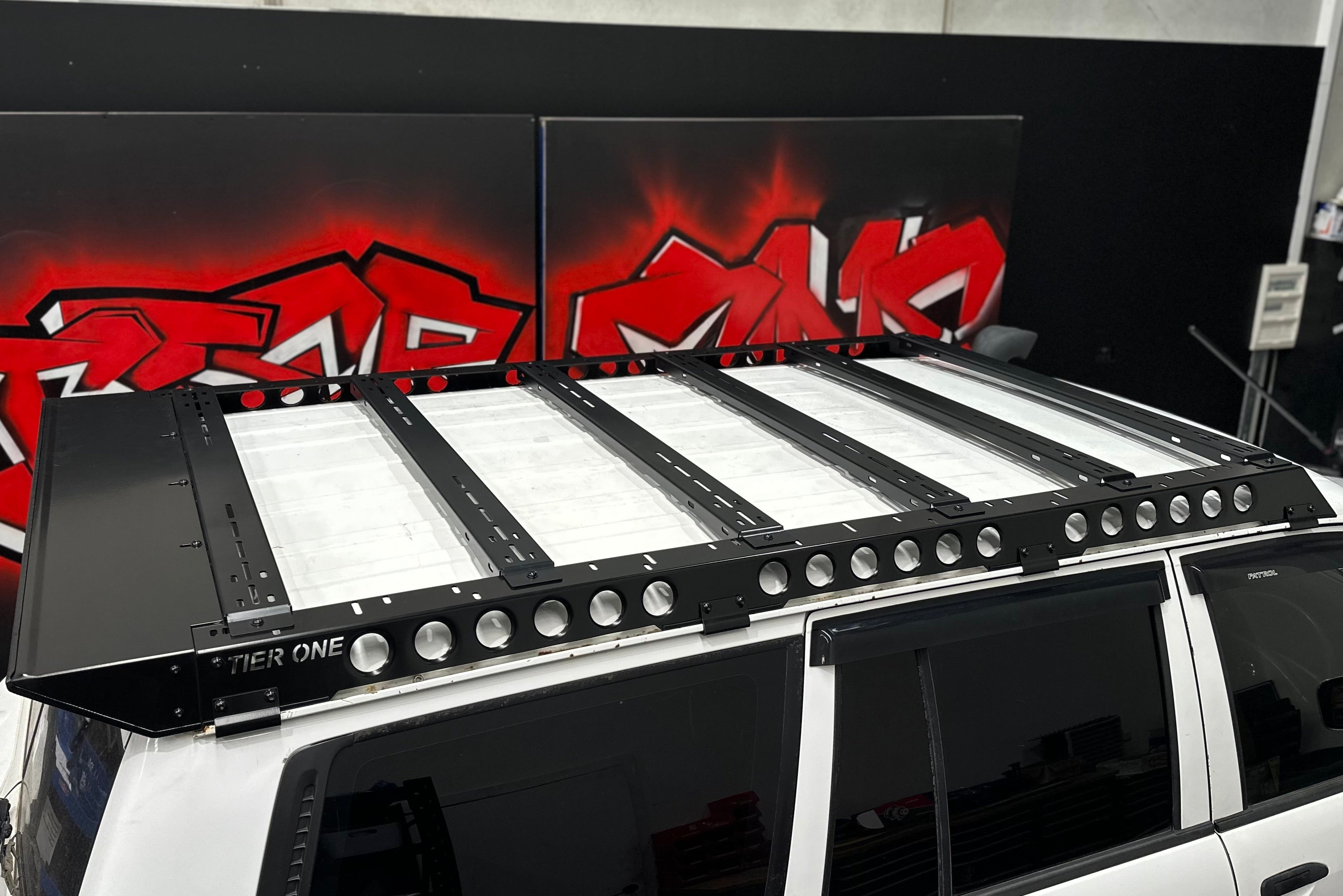 This is an image of a black Tier One Metalworks roof rack mounted on a white 4x4 SUV in a garage setting