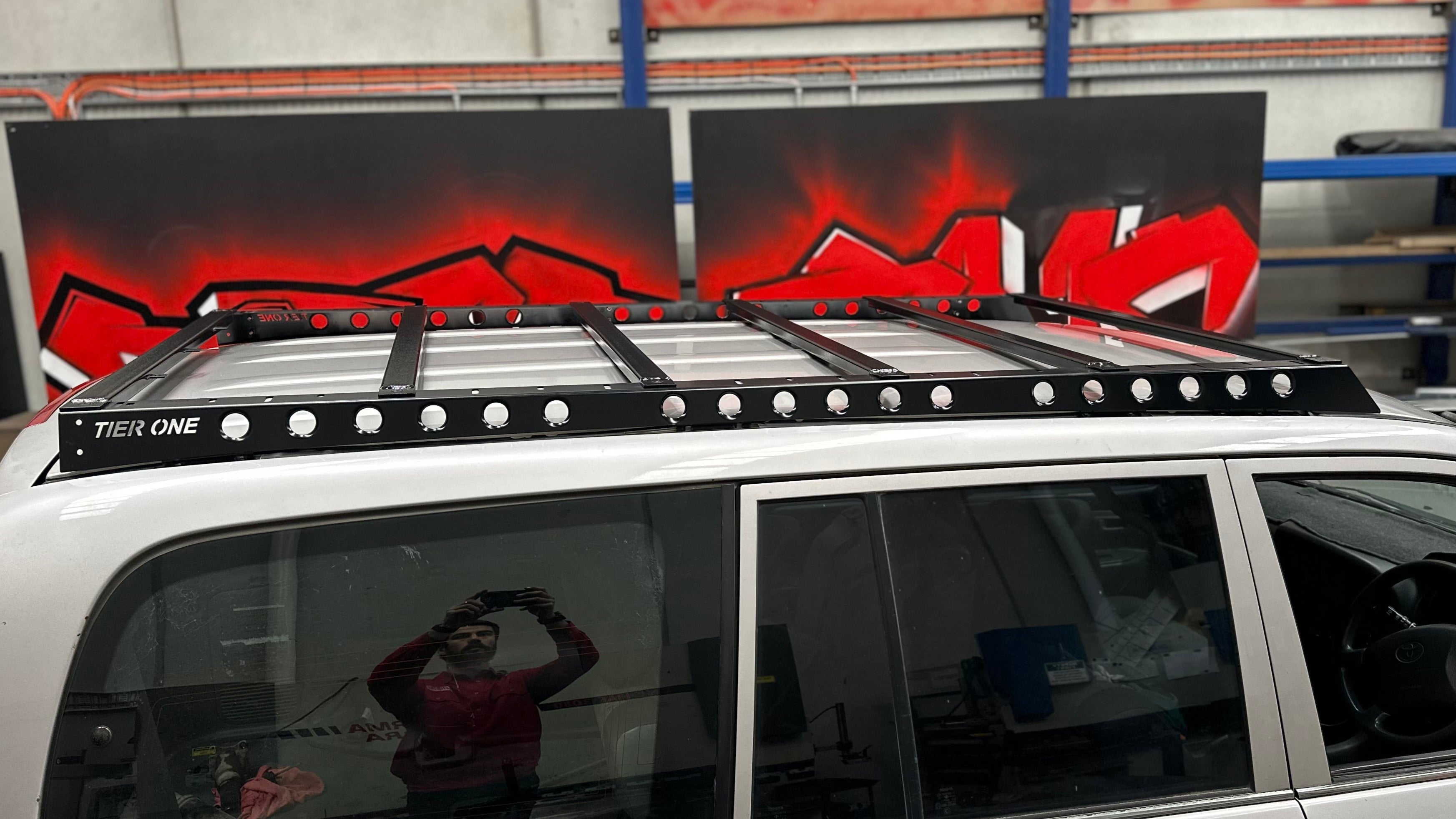 This is an image of a white 4x4 with a black Tier One Metalworks Roof Rack & Rails installed, shown indoors with red graffiti in background.