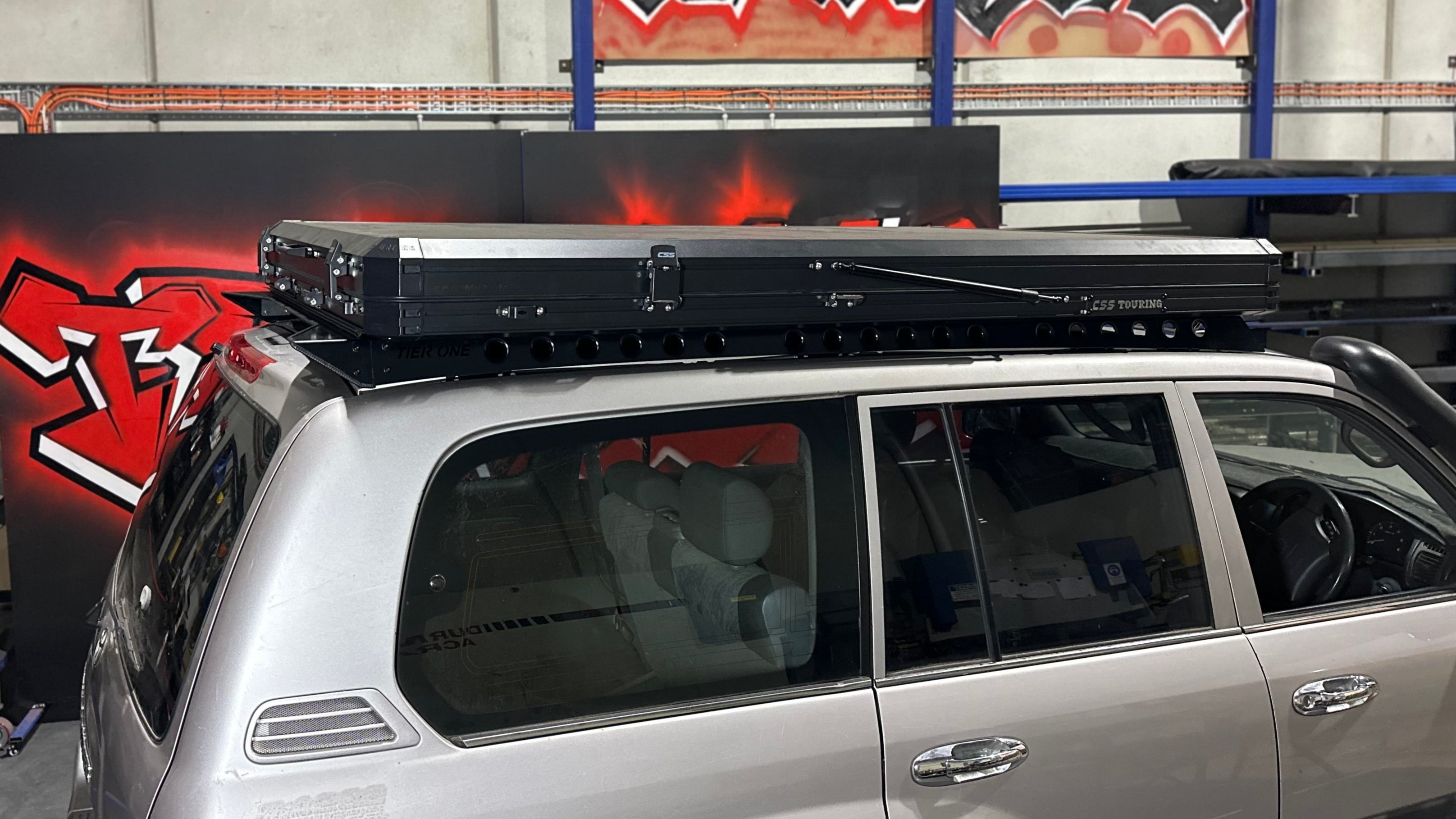 This is an image of a silver Toyota LandCruiser 200 series with a black Tier One Metalworks Roof Rack and CSS Touring rooftop tent in a workshop.