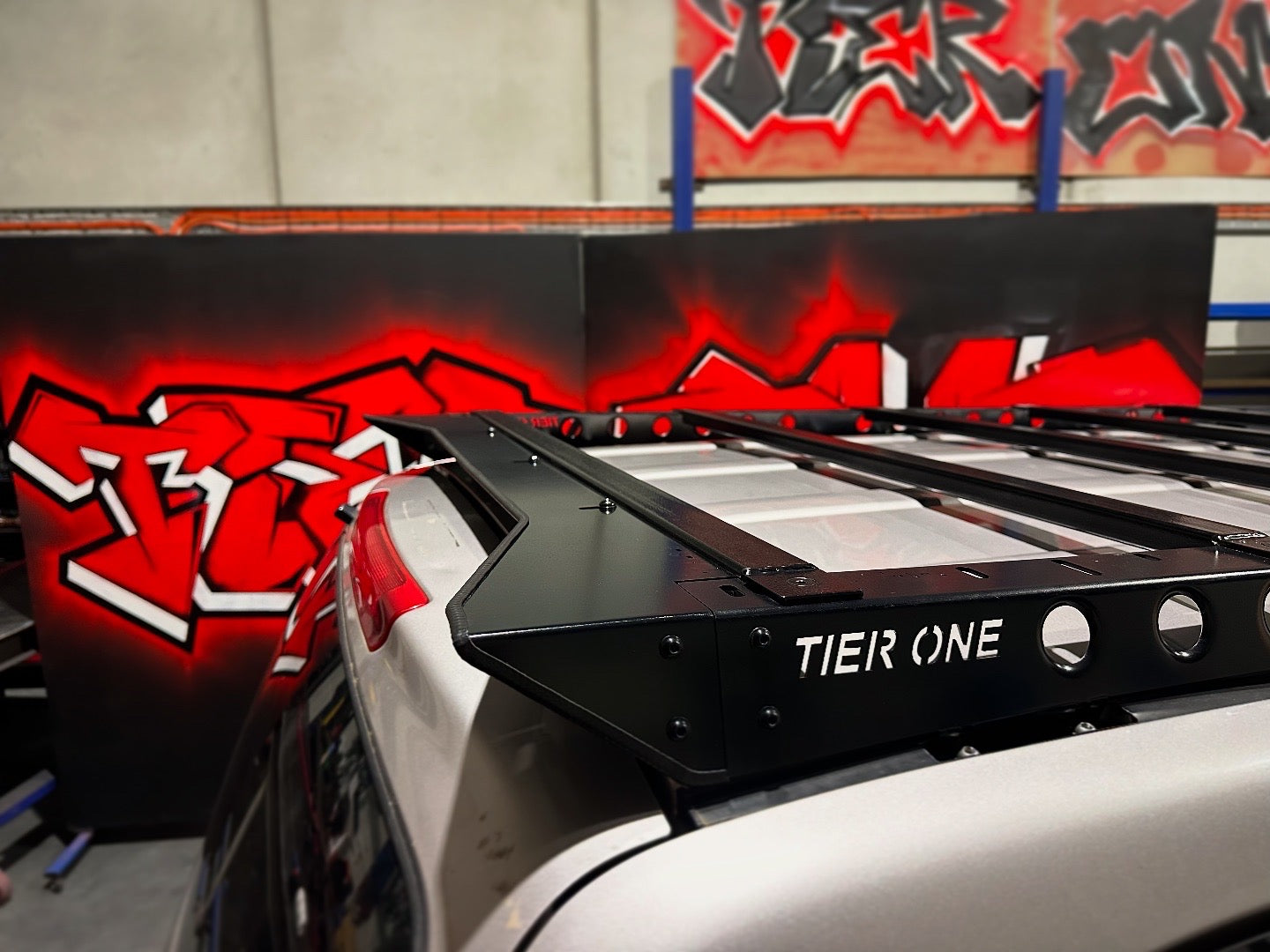This is an image of a black Tier One Metalworks roof rack fitted on a silver 4WD vehicle with red graffiti background