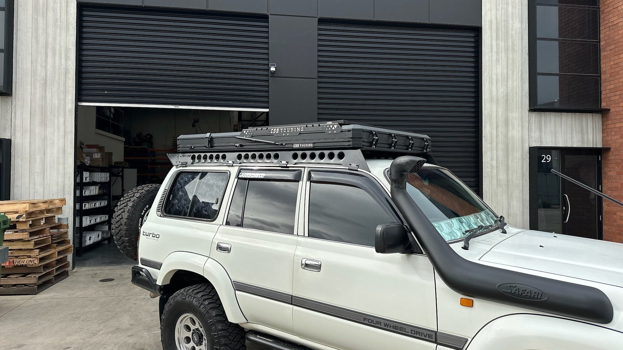 This is an image of a white 4WD SUV fitted with a Safari snorkel and a Tier One Metalworks roof rack with CSS Touring rooftop tent, parked outside an industrial garage.