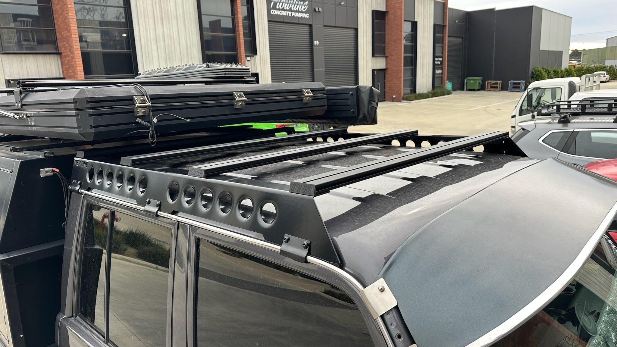 This is an image of a grey 4x4 vehicle featuring a Tier One Metalworks black roof rack and rails mounted on the roof in an industrial area