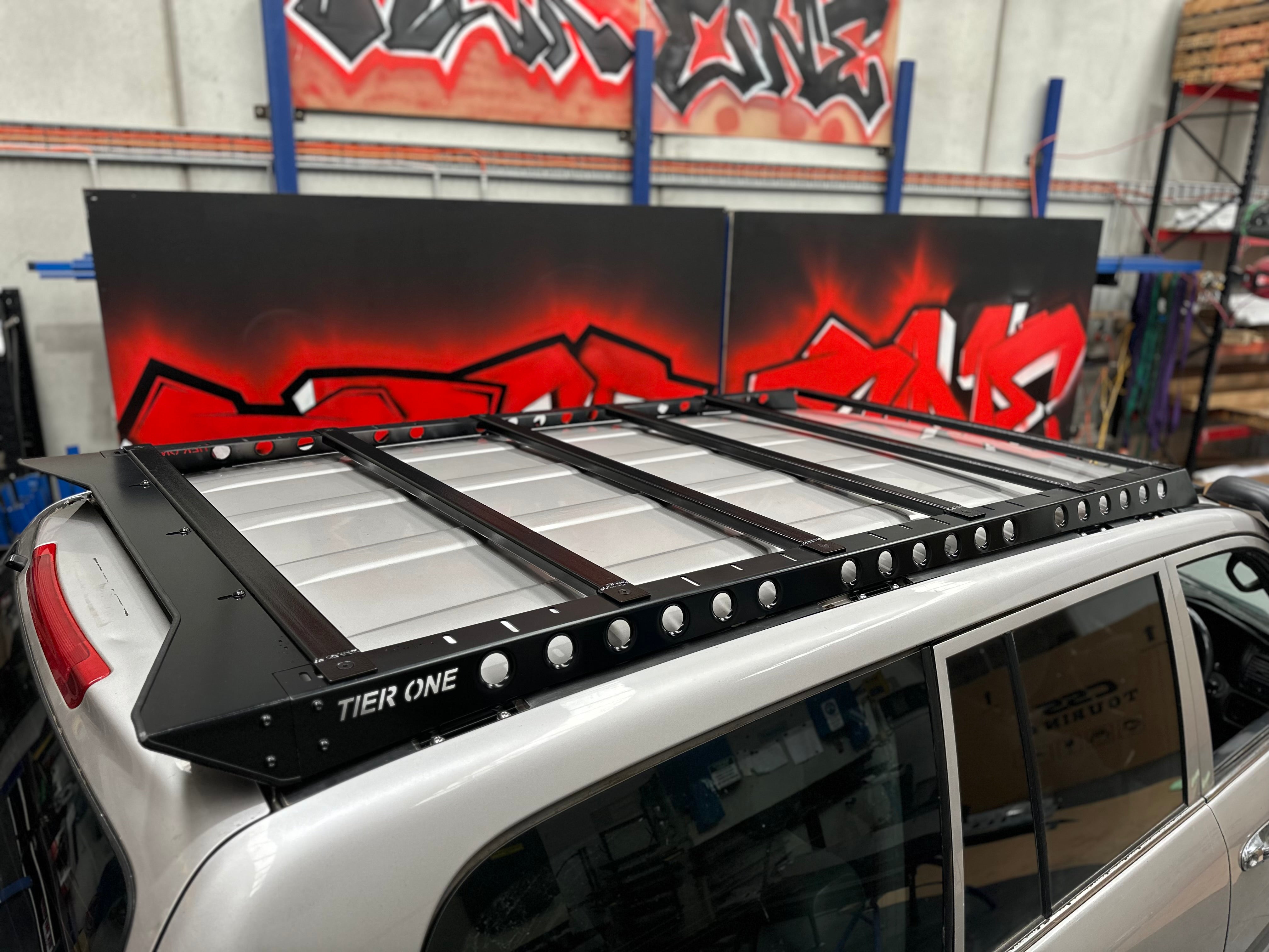 This is an image of a black Tier One Metalworks roof rack mounted on a silver 4x4 vehicle in a workshop with red graffiti background.