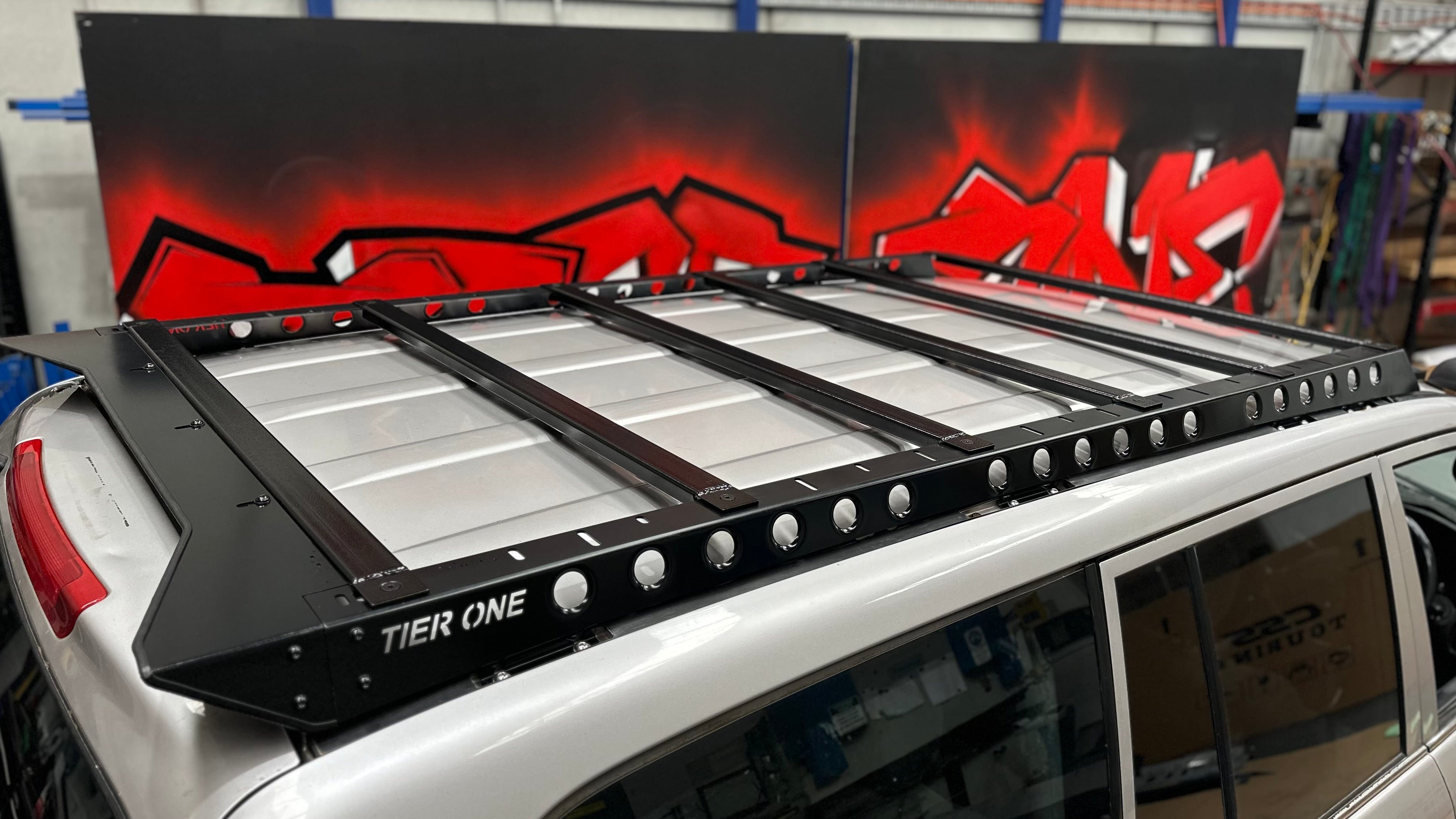 This is an image of a black Tier One Metalworks roof rack mounted on a silver 4x4 vehicle in a workshop with red graffiti background.