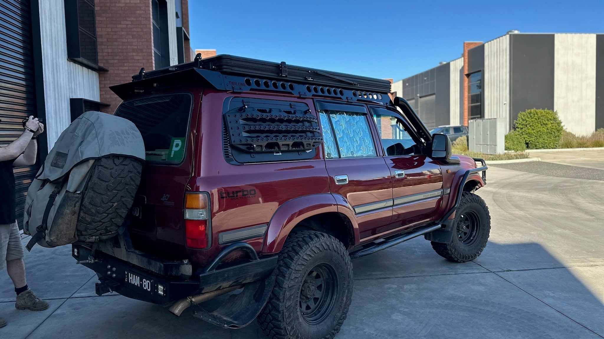 This is an image of a maroon Toyota Land Cruiser 80 Series with Tier One Metalworks black roof rack and side rails parked outside industrial buildings.