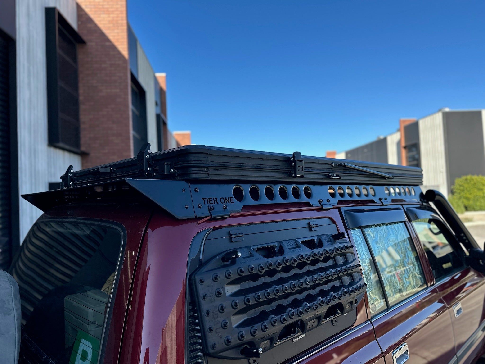 This is an image of a maroon 4WD fitted with a Tier One Metalworks black roof rack and side-mounted recovery tracks under clear blue sky