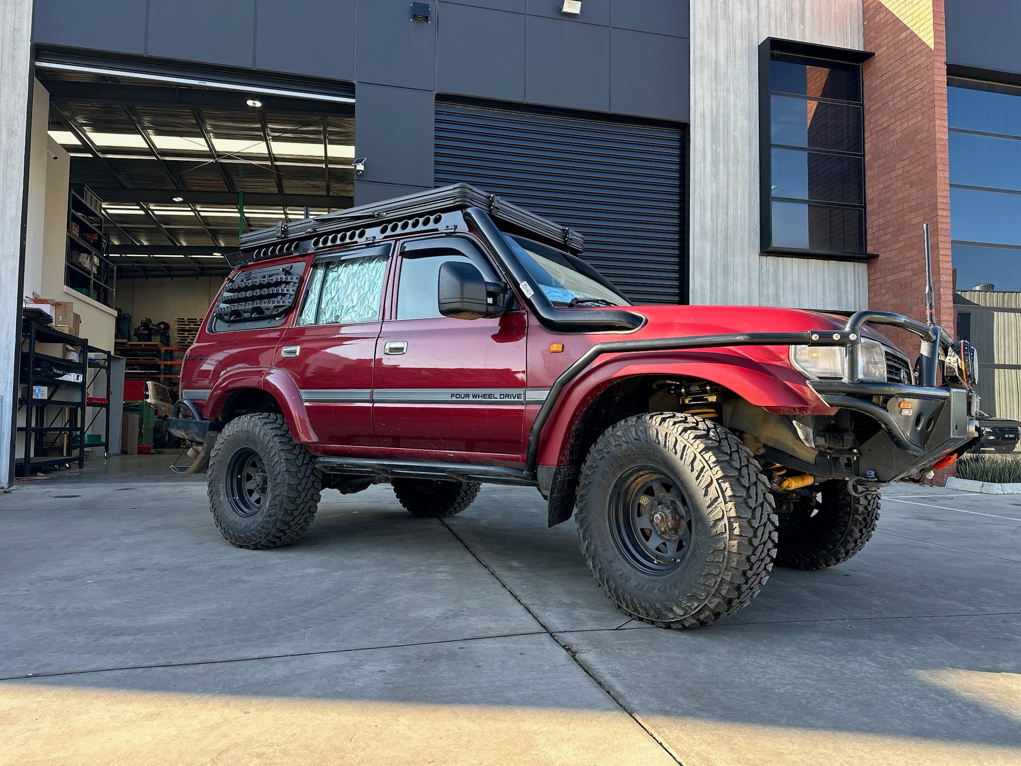 This is an image of a red Toyota Land Cruiser 80 Series with Tier One Metalworks black roof racks and snorkel in an industrial workshop setting