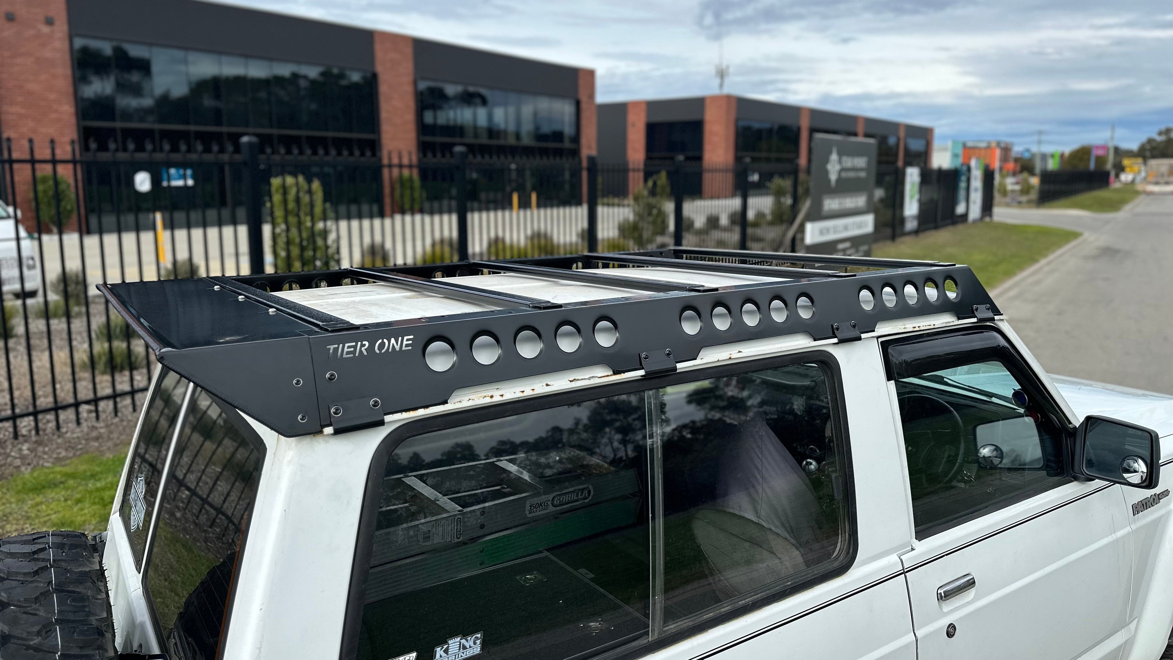 This is an image of a white Nissan Patrol 4x4 with a black Tier One Metalworks Roof Rack mounted on top in an industrial area.