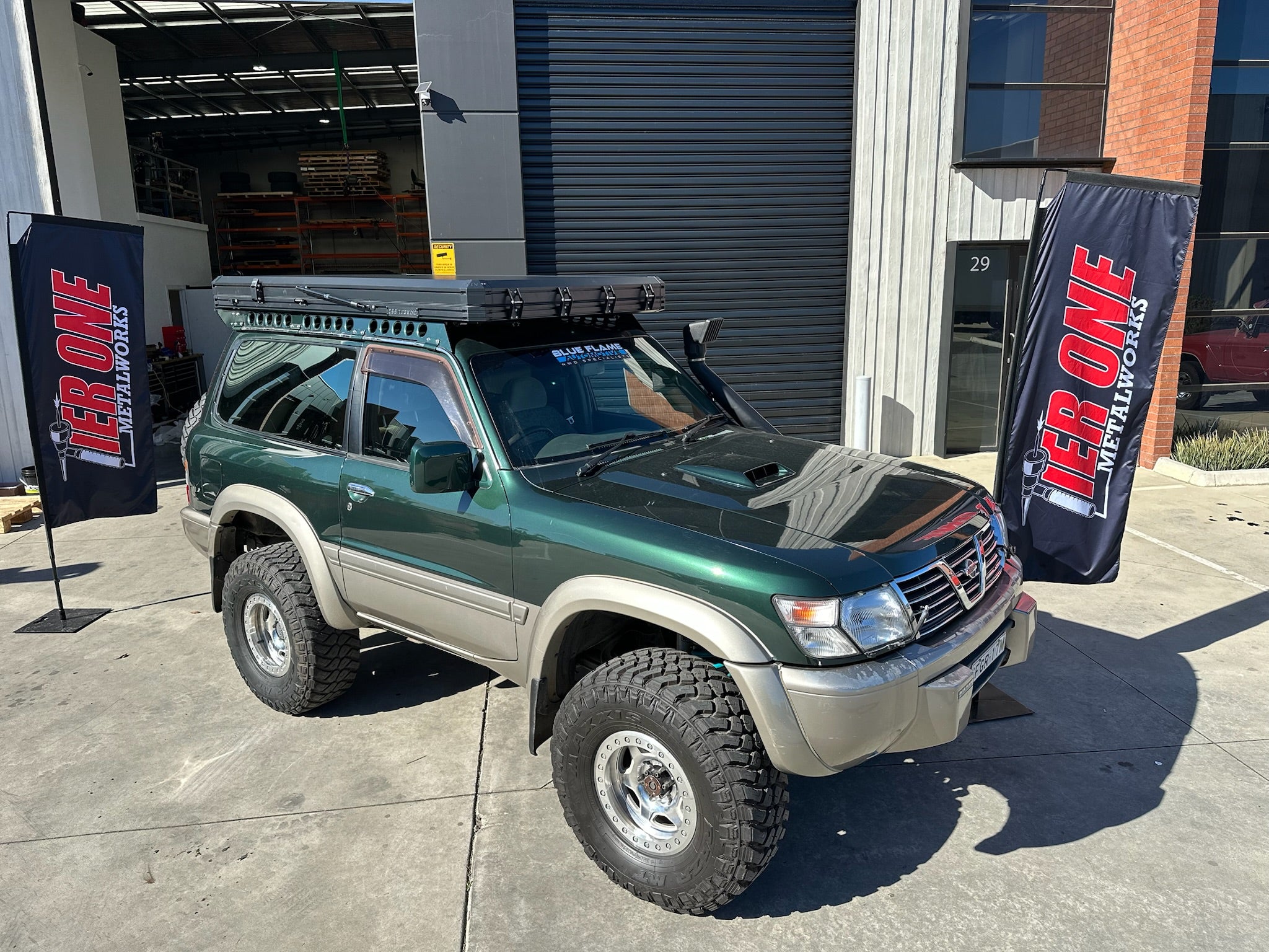 This is an image of a green and beige Nissan Patrol 4WD with Tier One Metalworks custom roof rack and snorkel parked outside a workshop.