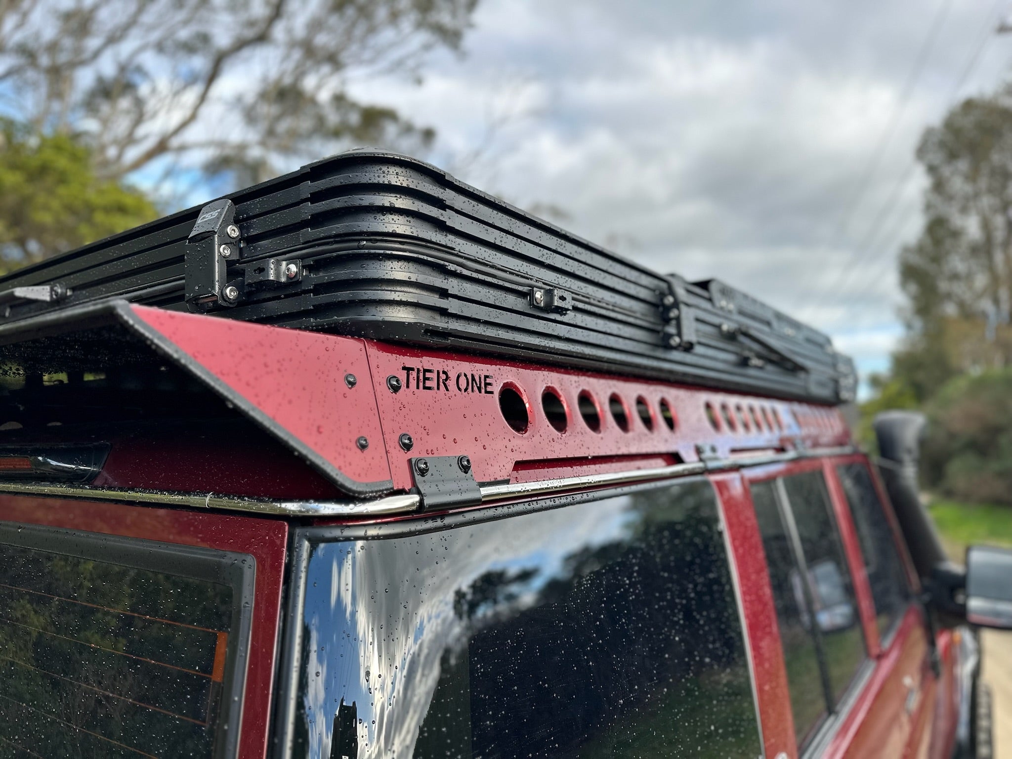 This is an image of a red 4x4 with a Tier One Metalworks black roof rack fitted on top on a rainy day