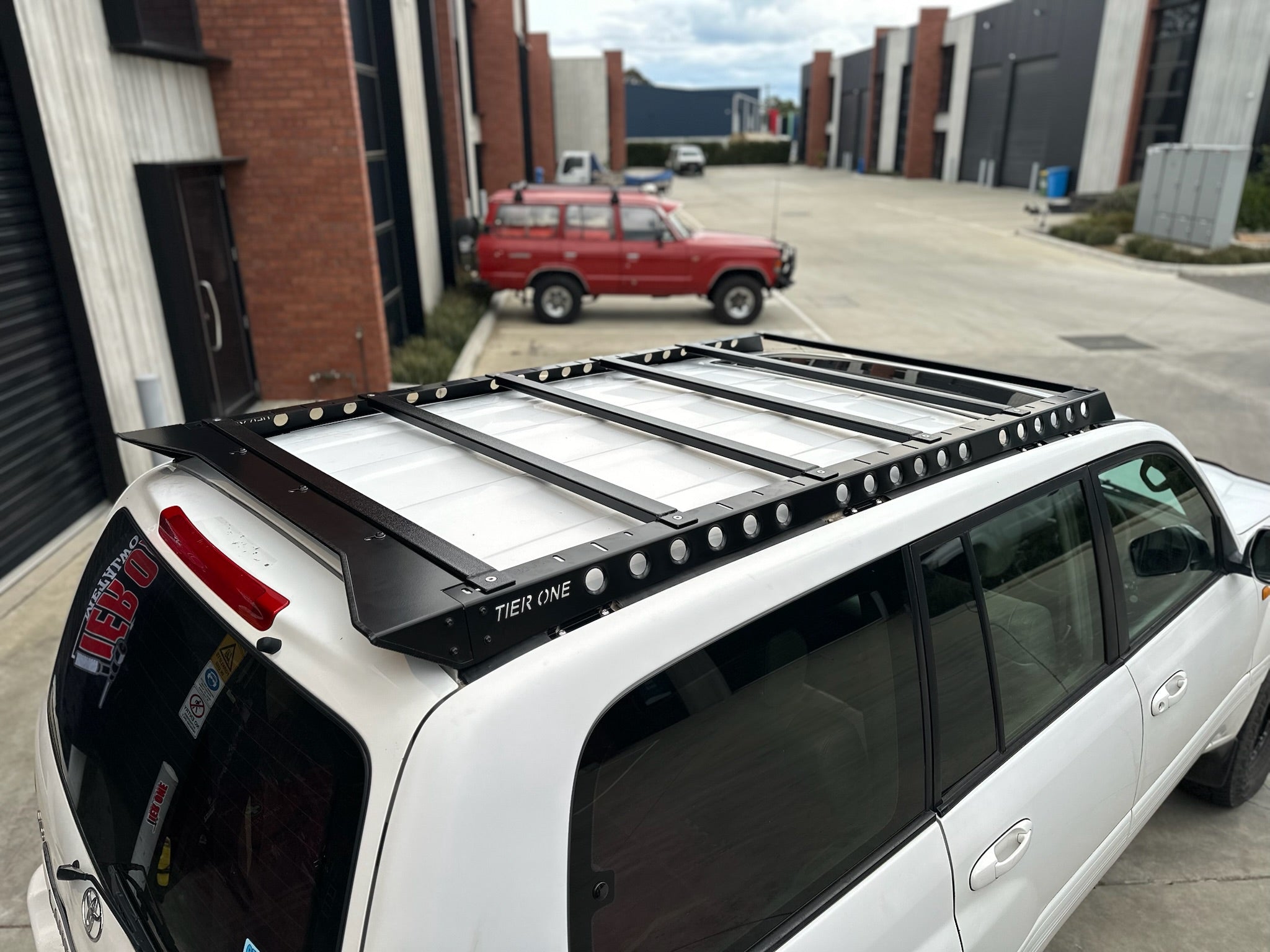 This is an image of a white Toyota 4x4 fitted with a black Tier One Metalworks roof rack in an industrial parking lot