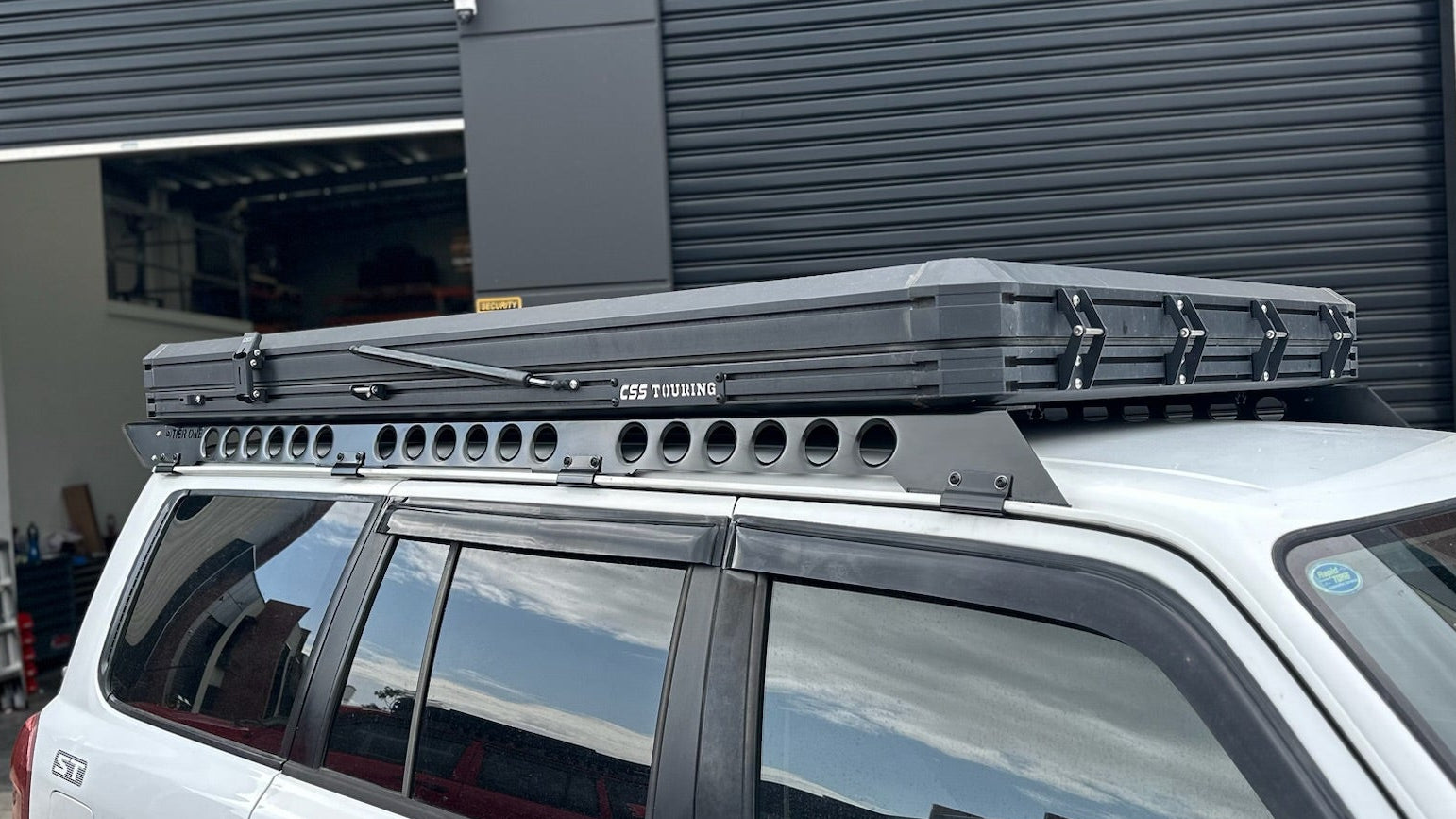 This is an image of a white Nissan Patrol with black Tier One Metalworks Roof Racks & CSS Touring rooftop tent in a workshop