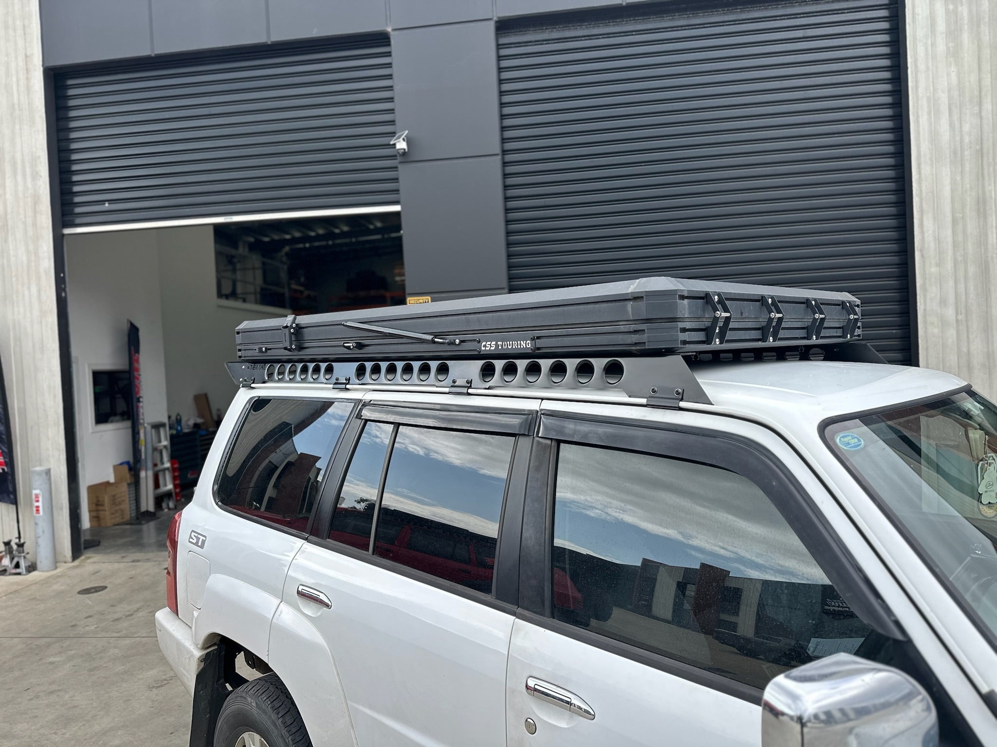 This is an image of a white Nissan Patrol with black Tier One Metalworks Roof Racks & CSS Touring rooftop tent in a workshop