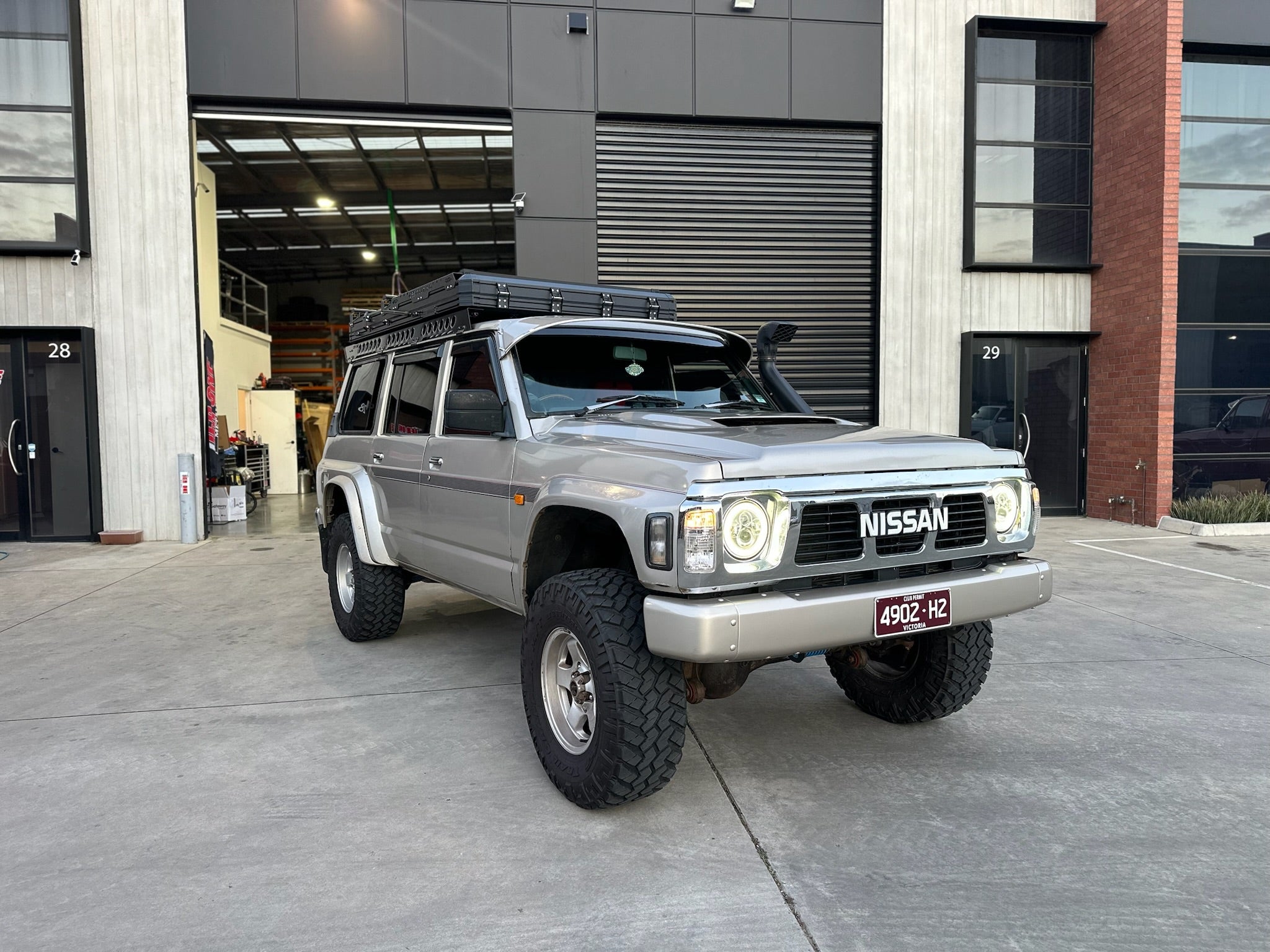 This is an image of a silver Nissan Patrol 4WD fitted with Tier One Metalworks custom roof rack and snorkel, parked outside a modern workshop