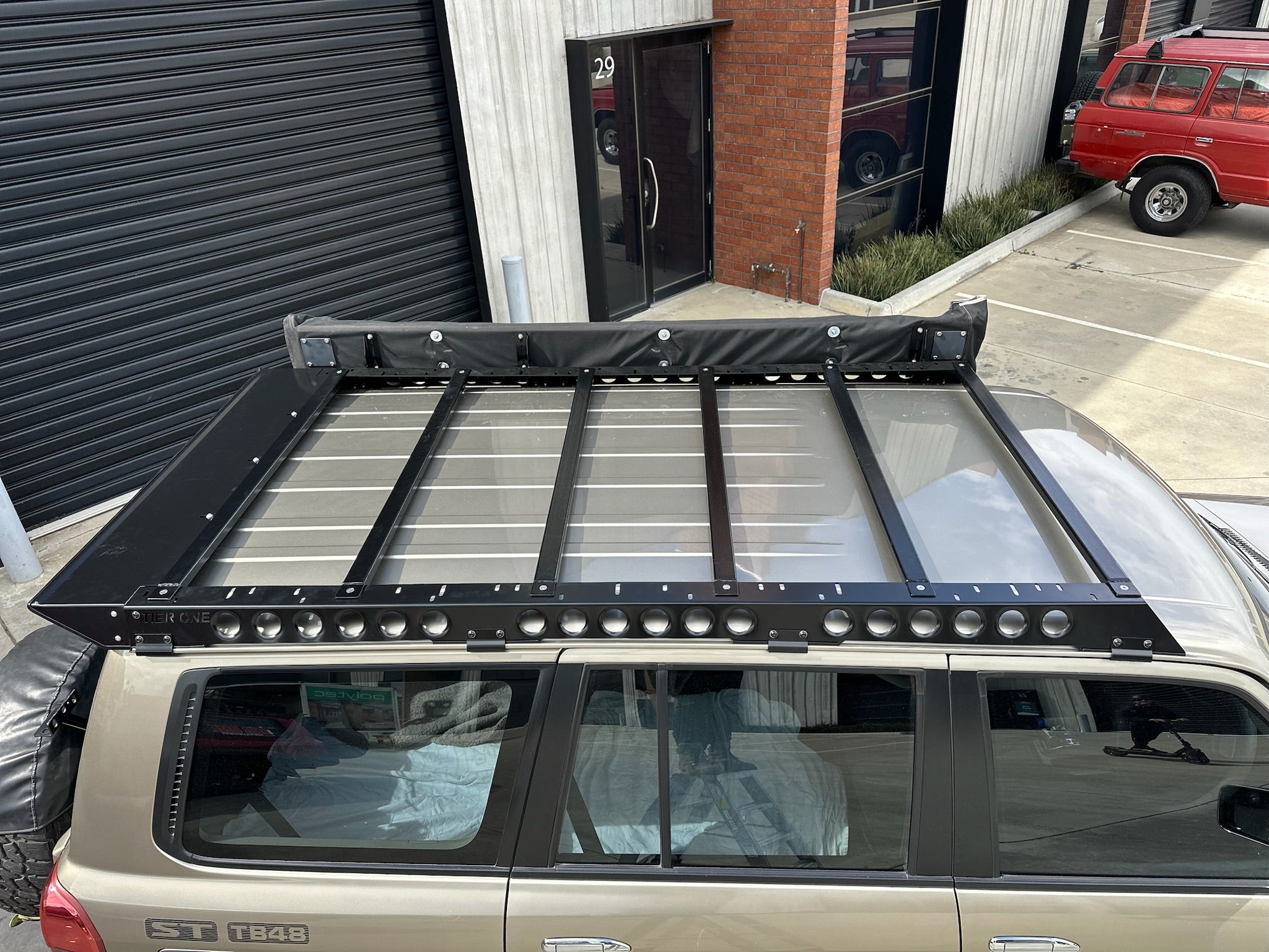 This is an image of a beige Toyota Land Cruiser Prado 120 Series with a black Tier One Metalworks Roof Rack mounted on top, parked outside a commercial building.