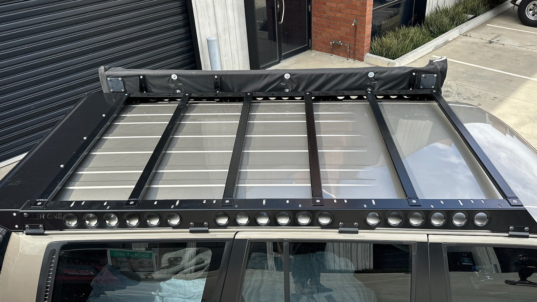 This is an image of a beige Toyota Land Cruiser Prado 120 Series with a black Tier One Metalworks Roof Rack mounted on top, parked outside a commercial building.