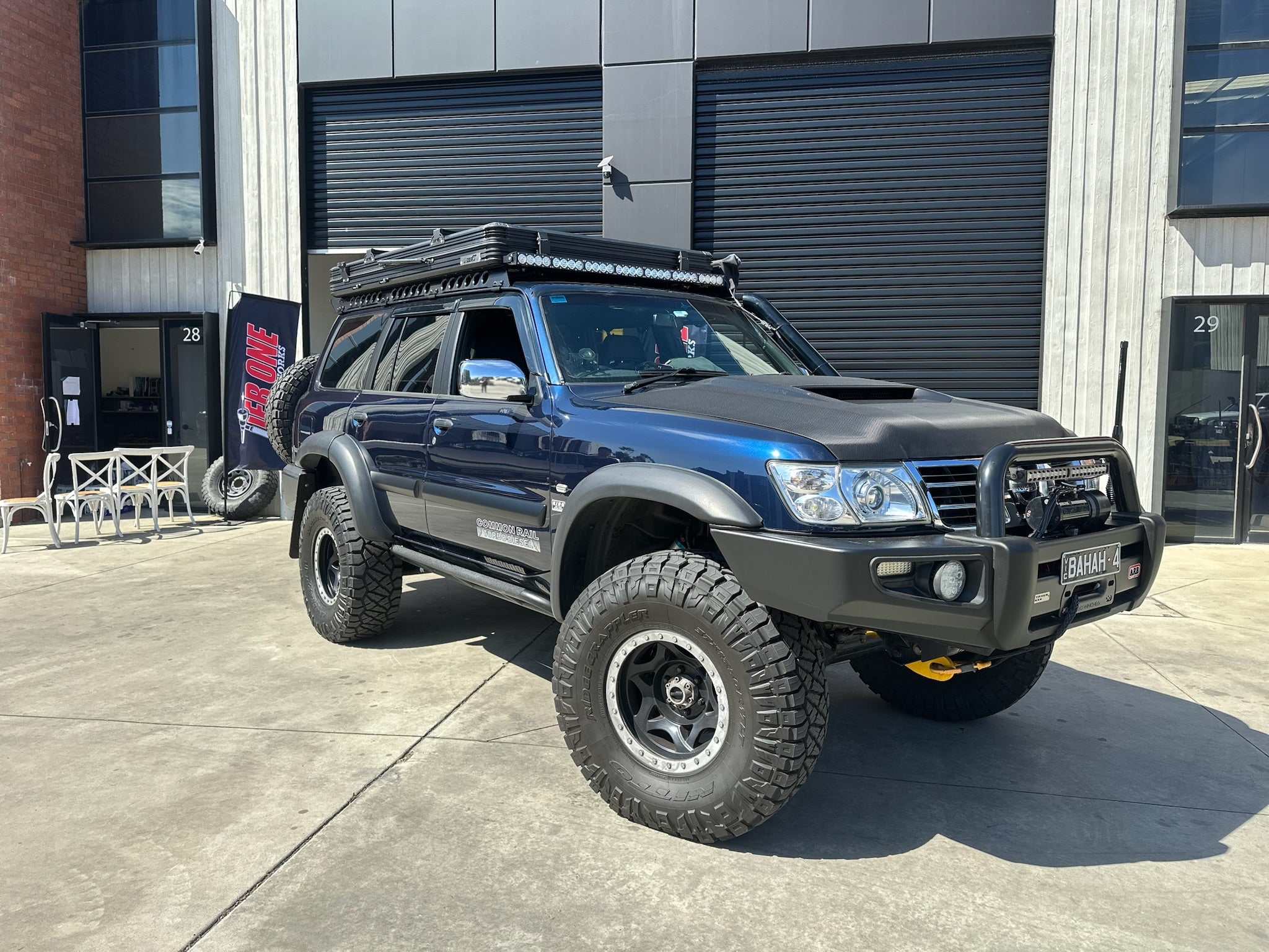 This is an image of a blue Toyota LandCruiser 100 Series with Tier One Metalworks roof racks and custom bull bar in an industrial setting.