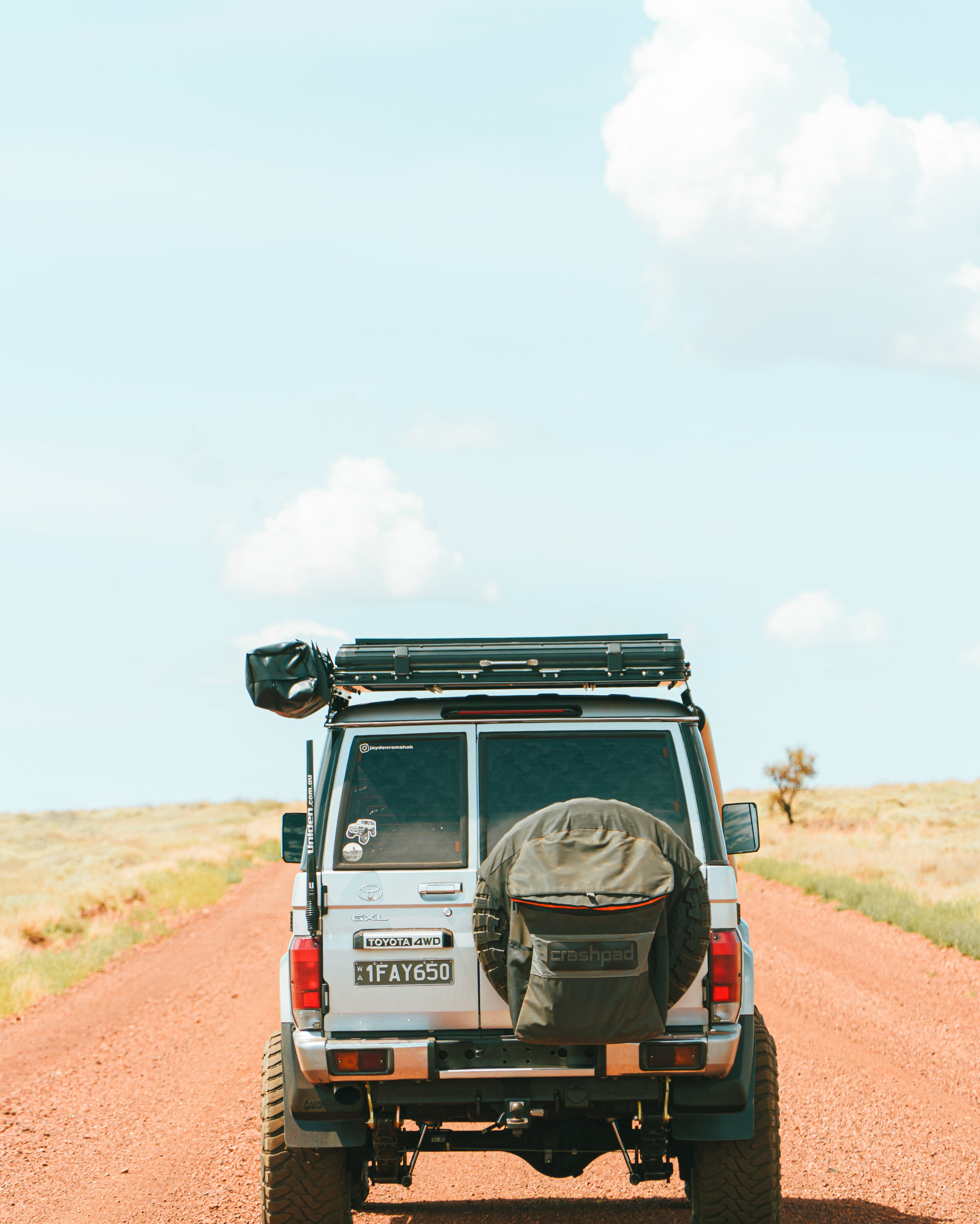 This is an image of a white Toyota Landcruiser 76 Series driving on a red dirt road in outback Australia featuring a black Tier One Metalworks roof rack with rear wing and a spare tyre cover