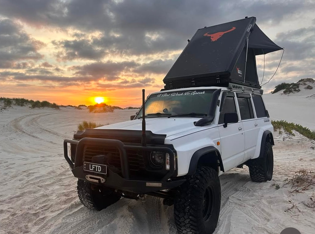 This is an image of a white Nissan Patrol 4x4 with a bull bar, snorkel, and rooftop tent at sunset on sandy terrain, featuring Tier One Metalworks Roof Racks & Rails.