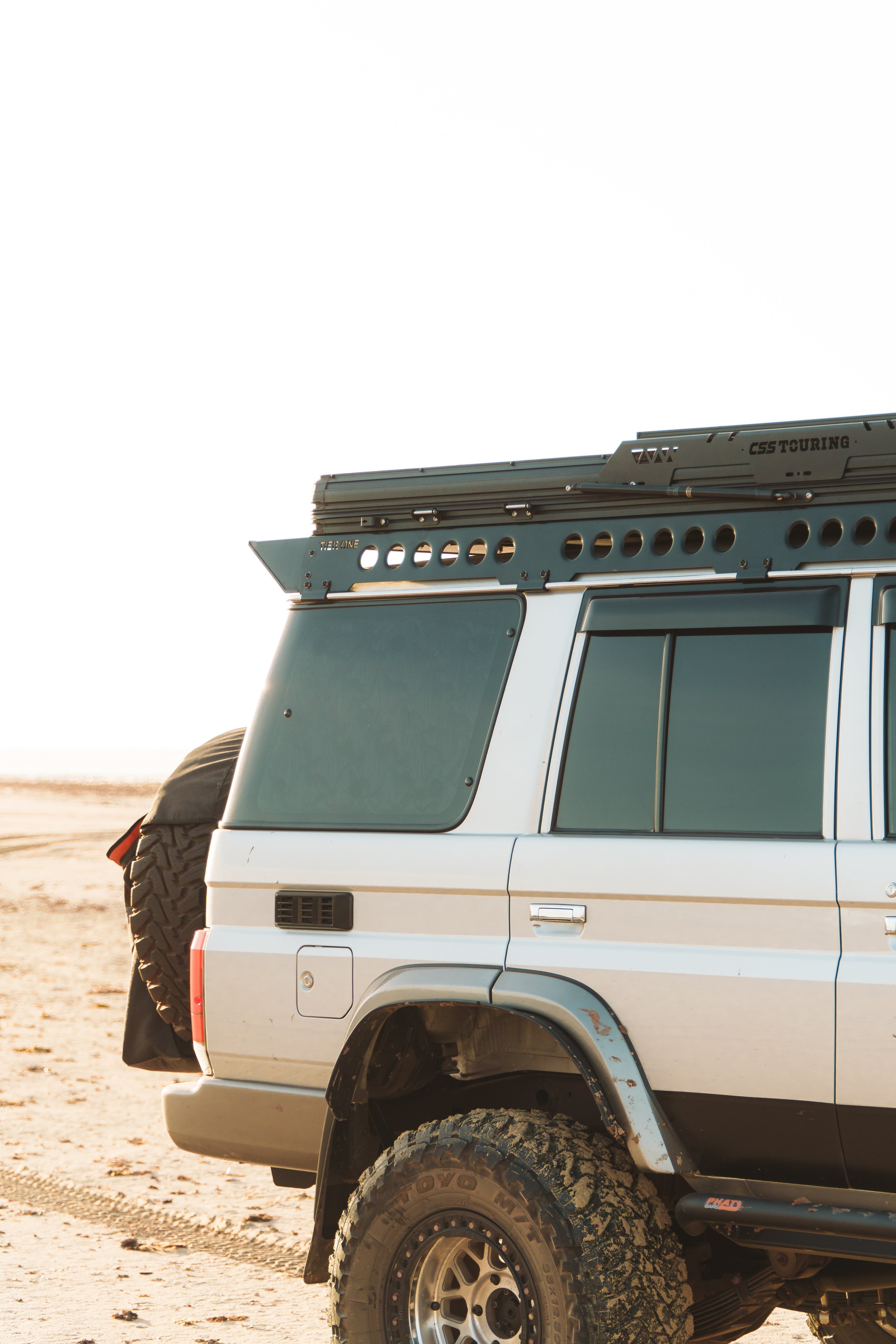 This is an image of a white 4x4 vehicle equipped with Tier One Metalworks Roof Racks & Rails and a CSS Touring rooftop tent on sandy terrain.